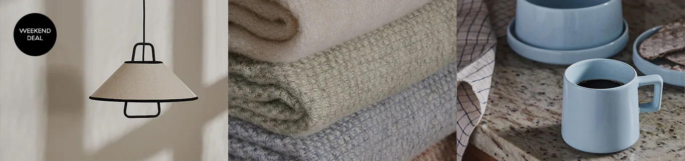 Een weekenddeal met een crèmekleurige hanglamp met zwarte rand, een stapel fleecedekens in beige, groen en blauw, en een lichtblauwe mok met koffie.