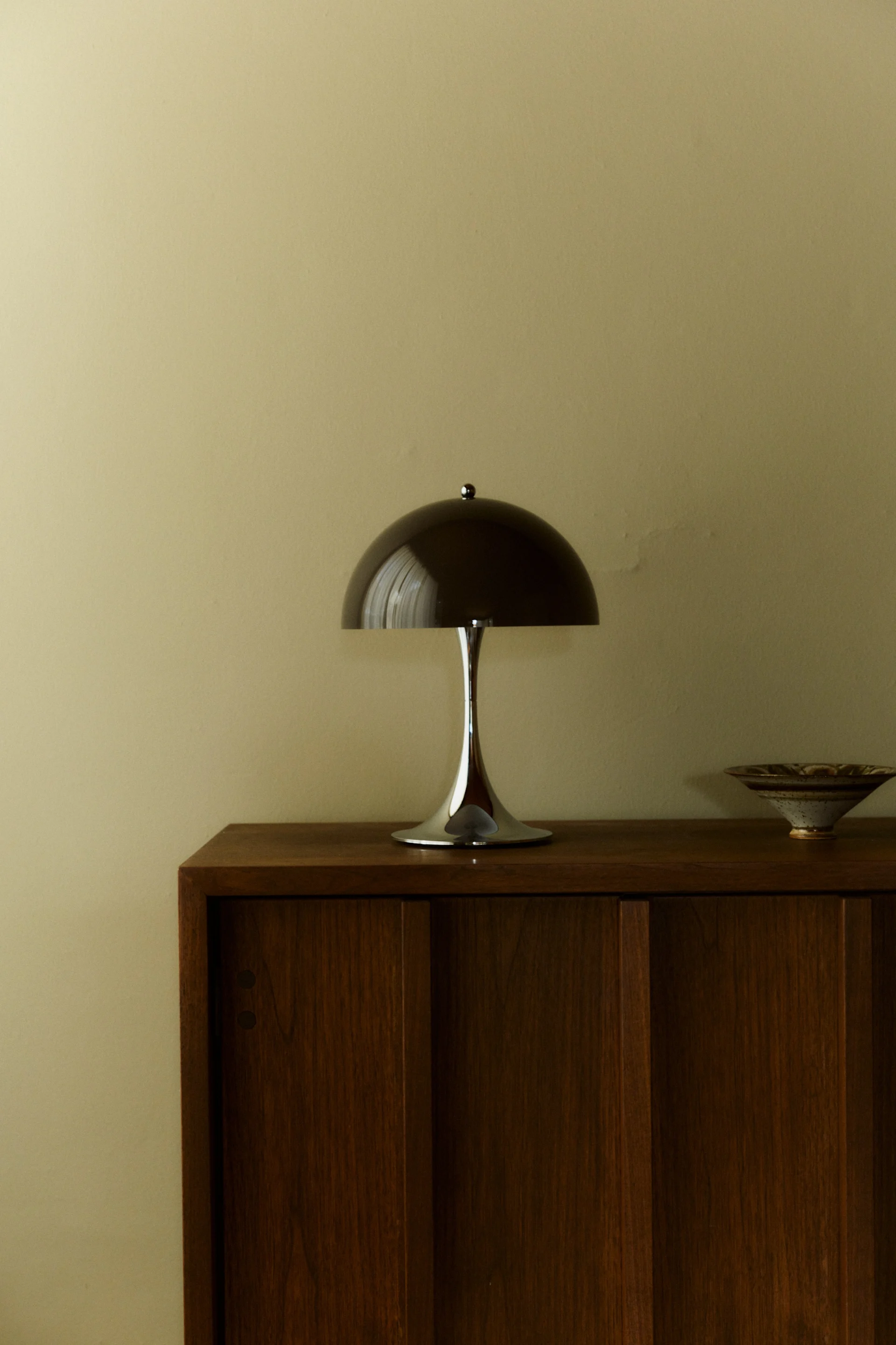 Een slanke, moderne champignon tafellamp met een donkere koepelvormige kap en chromen voet staat op een houten kast naast een keramische kom.