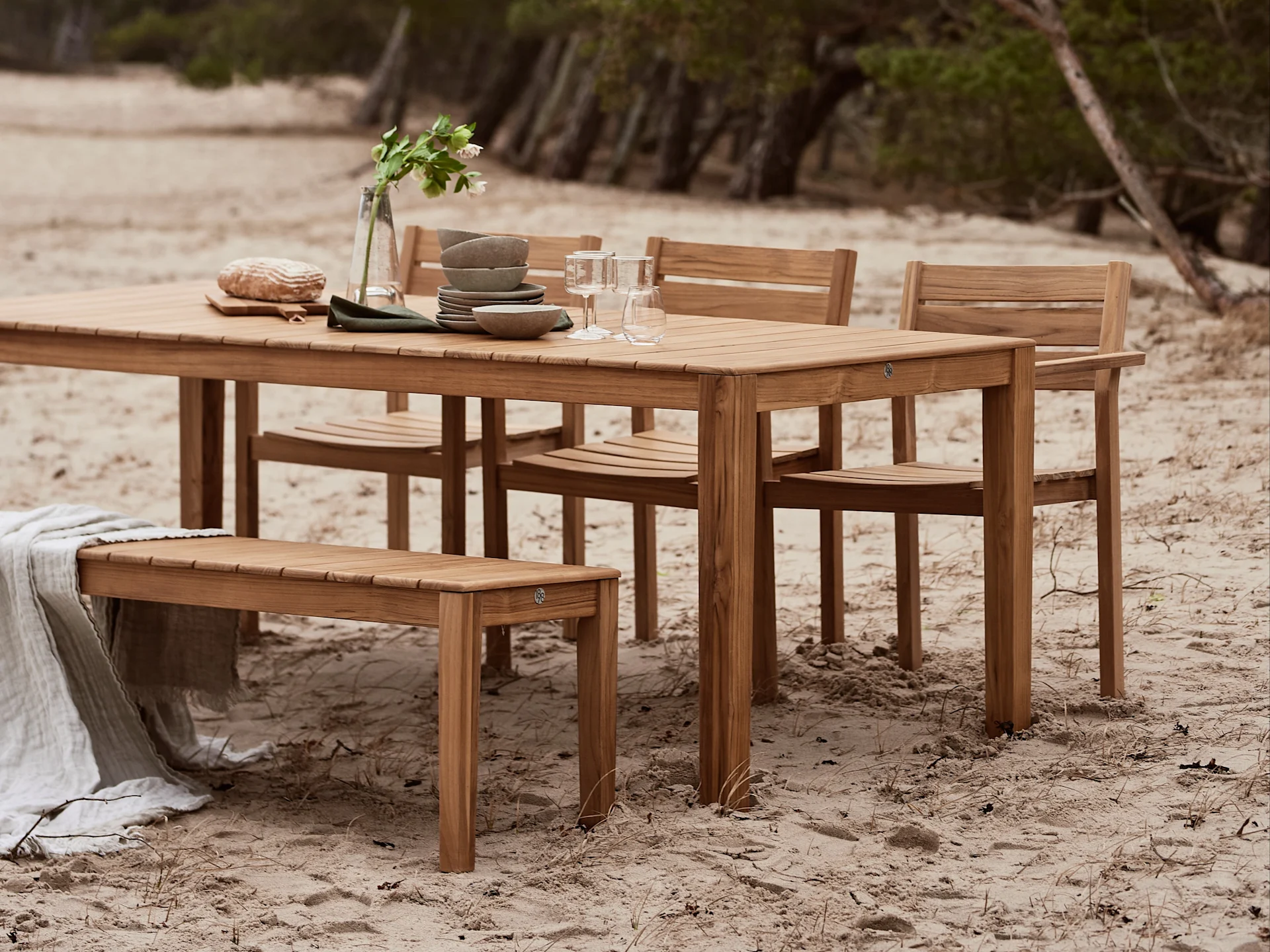 Houten buiten eetset met tafel, stoelen en bank, gestyled met servies en een deken op een zandstrand.