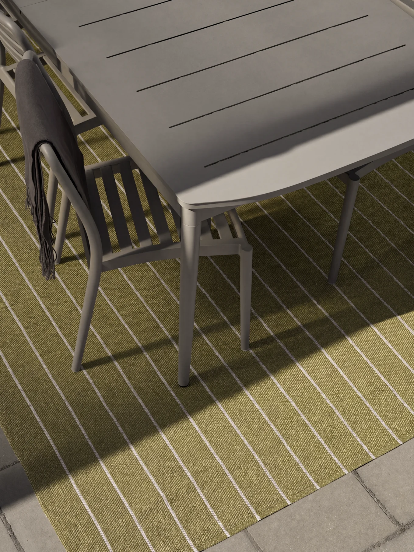 Grijze tuineettafel en stoelen op een groen-wit gestreept kleed. Een donkergrijze deken is over een stoel gedrapeerd.