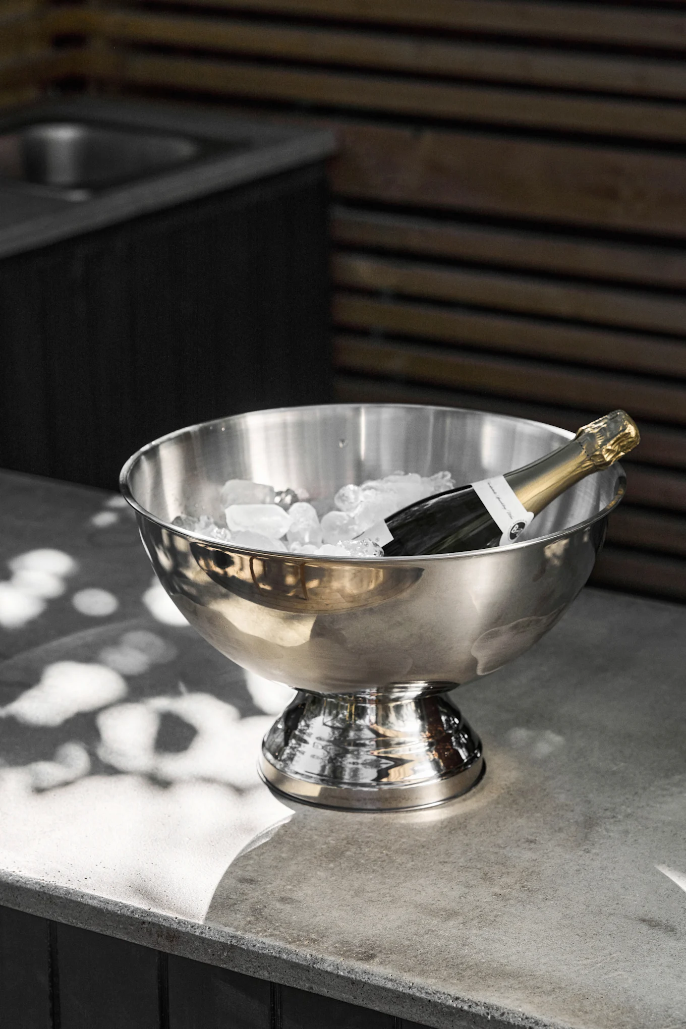 Een champagnefles rust in een roestvrijstalen ijsemmer gevuld met ijsblokjes op een grijze betonnen aanrecht, met gespikkeld zonlicht.