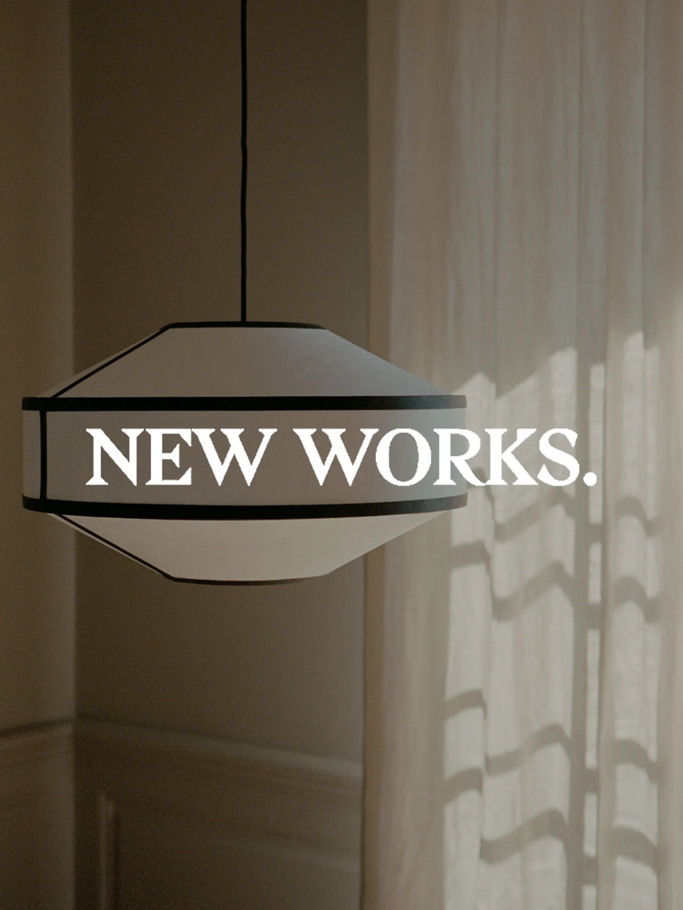 Een brede, wit- en donkergerande hanglamp hangt, met de tekst 'NEW WORKS.' erop. Zonlicht werpt schaduwen op een gordijn.