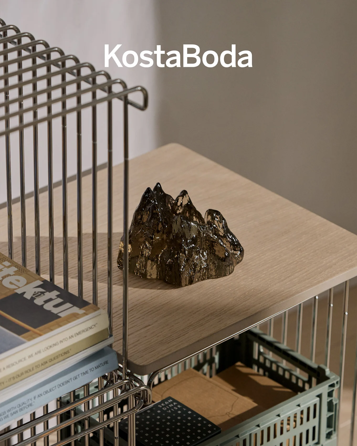 Merknaam KostaBoda boven een donker, transparant, bergvormig glasbeeld op een lichte houten plank.
