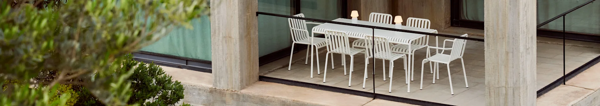Een buiten-eetset met een witte lamellentafel en zes bijpassende stoelen, verlicht door drie draagbare lampen, op een betonnen terras met planten.