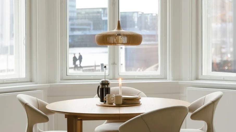 Ronde houten eettafel met vier lichte stoelen, een houten lamellen hanglamp, cafetière, mokken en borden erop.