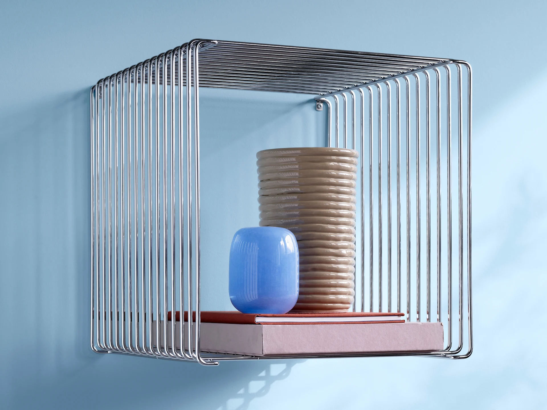 Een chromen draadrek met een hoge, geribbelde beige vaas, een kleine blauwe vaas en twee boeken aan een lichtblauwe muur.