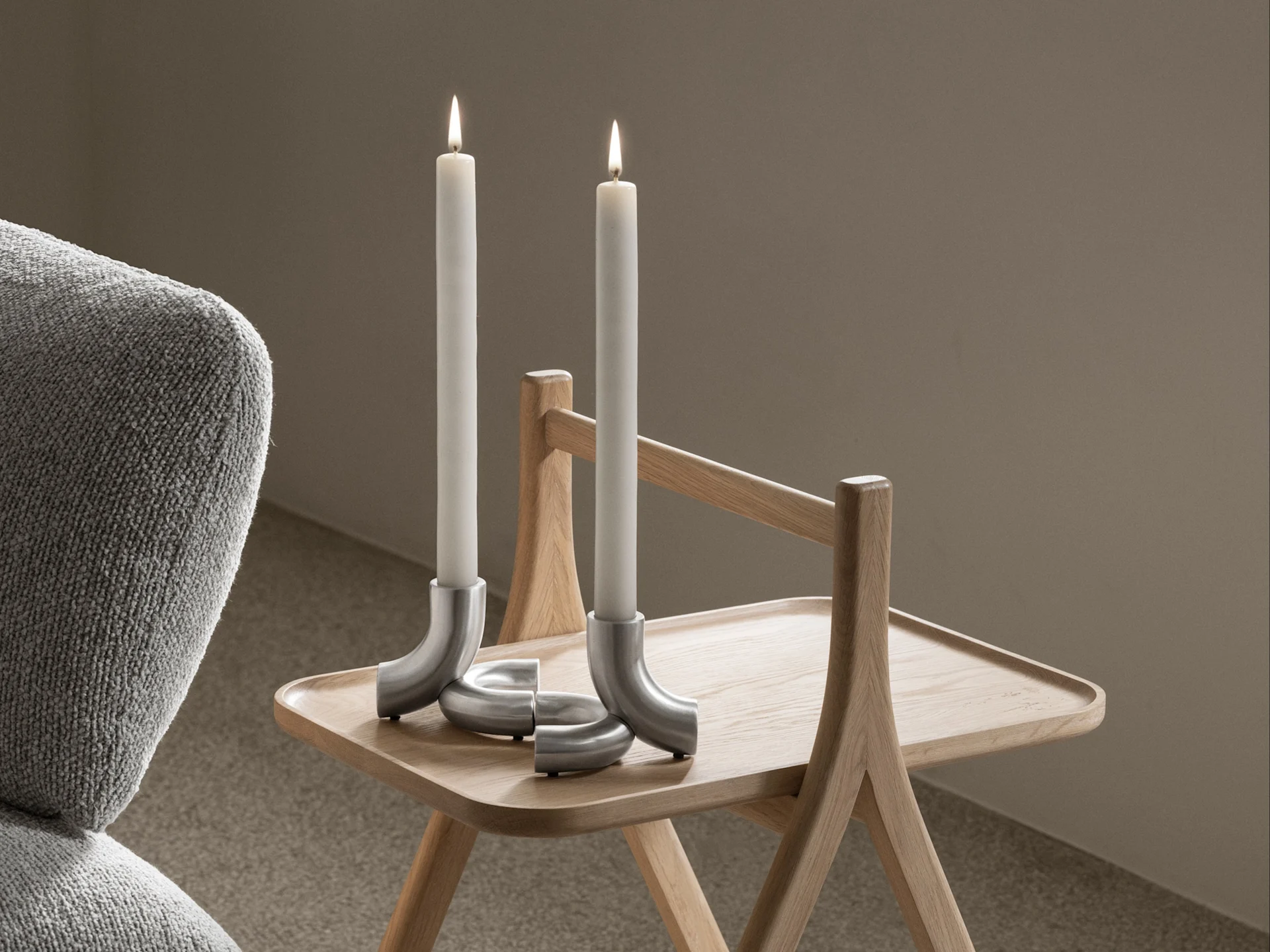 Twee brandende witte kaarsen in gebogen zilveren kandelaars op een lichte houten bijzettafel, met een grijze fauteuil links.