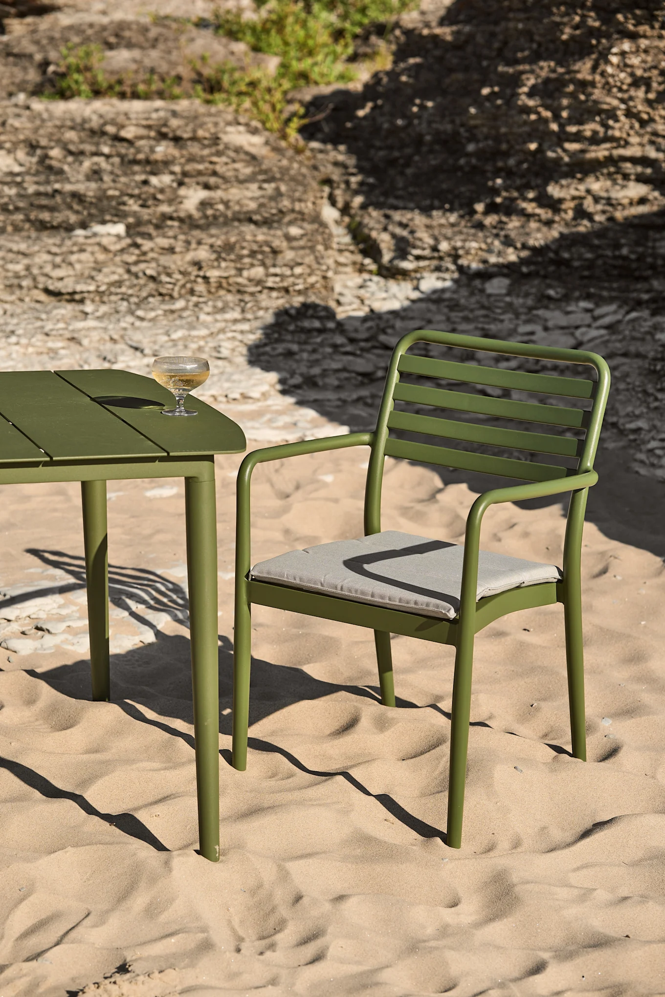 Groene metalen buitentafel en stoel op een zandstrand met een glas witte wijn op de tafel.
