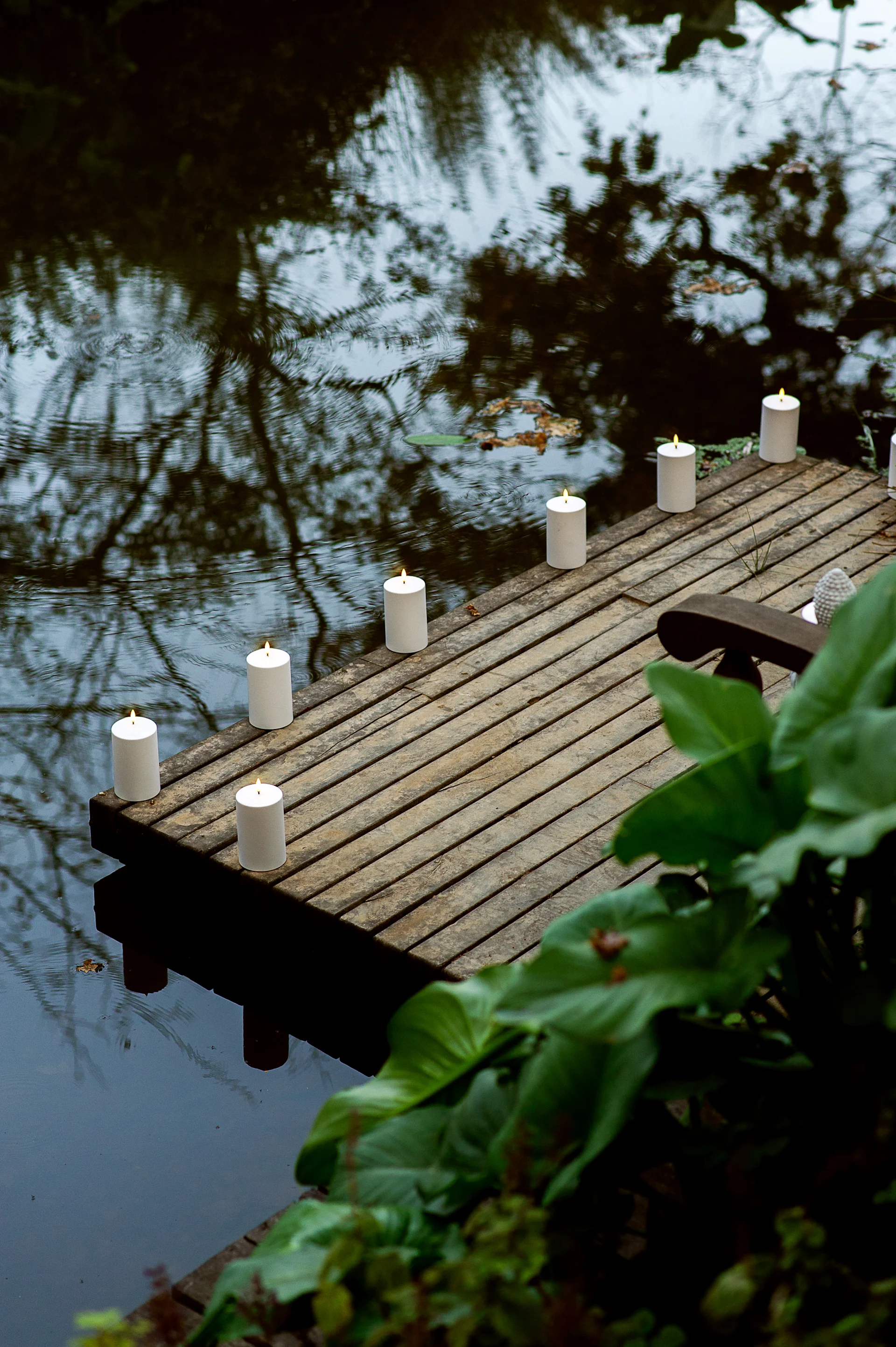 Verlichte witte pilaarkaarsen langs een houten steiger over een kalme vijver, met reflecties en groen blad.