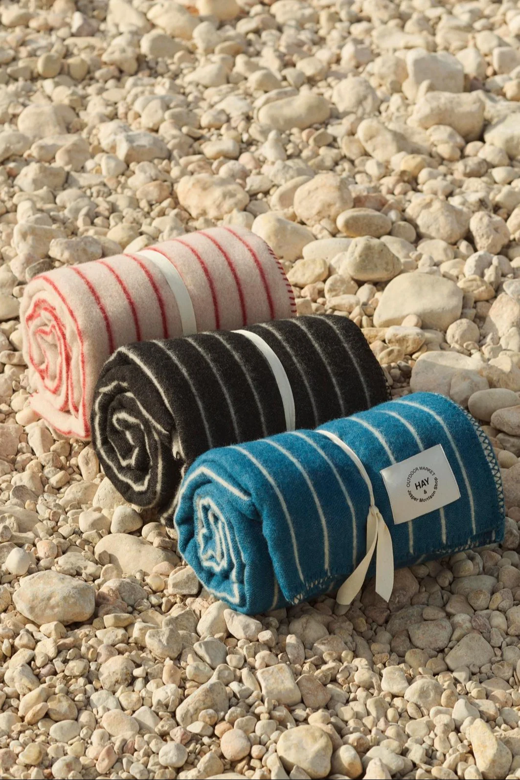 Drie opgerolde gestreepte dekens in roze, zwart en blauw, vastgebonden met linten op een kiezelstrand.