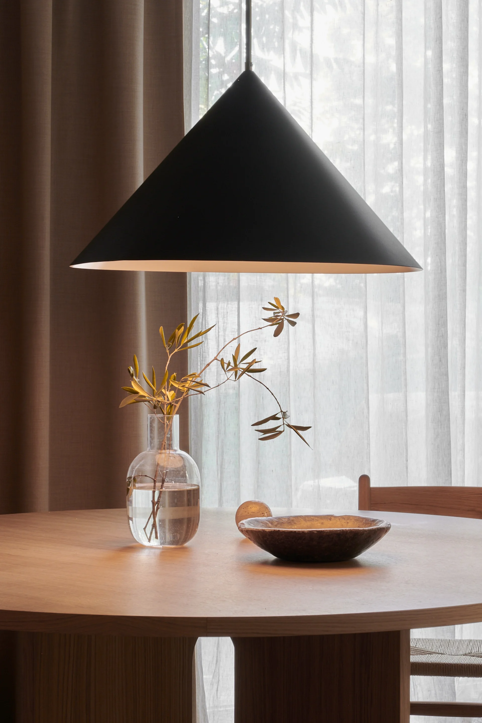 Ronde houten eettafel met een zwarte conische hanglamp erboven, voorzien van een heldere vaas met takken en een donkere houten kom.