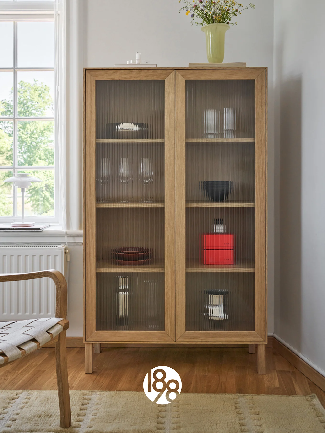 Een lichte houten kast met geribbelde glazen deuren met servies en glazen, bovenaan een vaas met wilde bloemen. Een stoel en raam bevinden zich links.