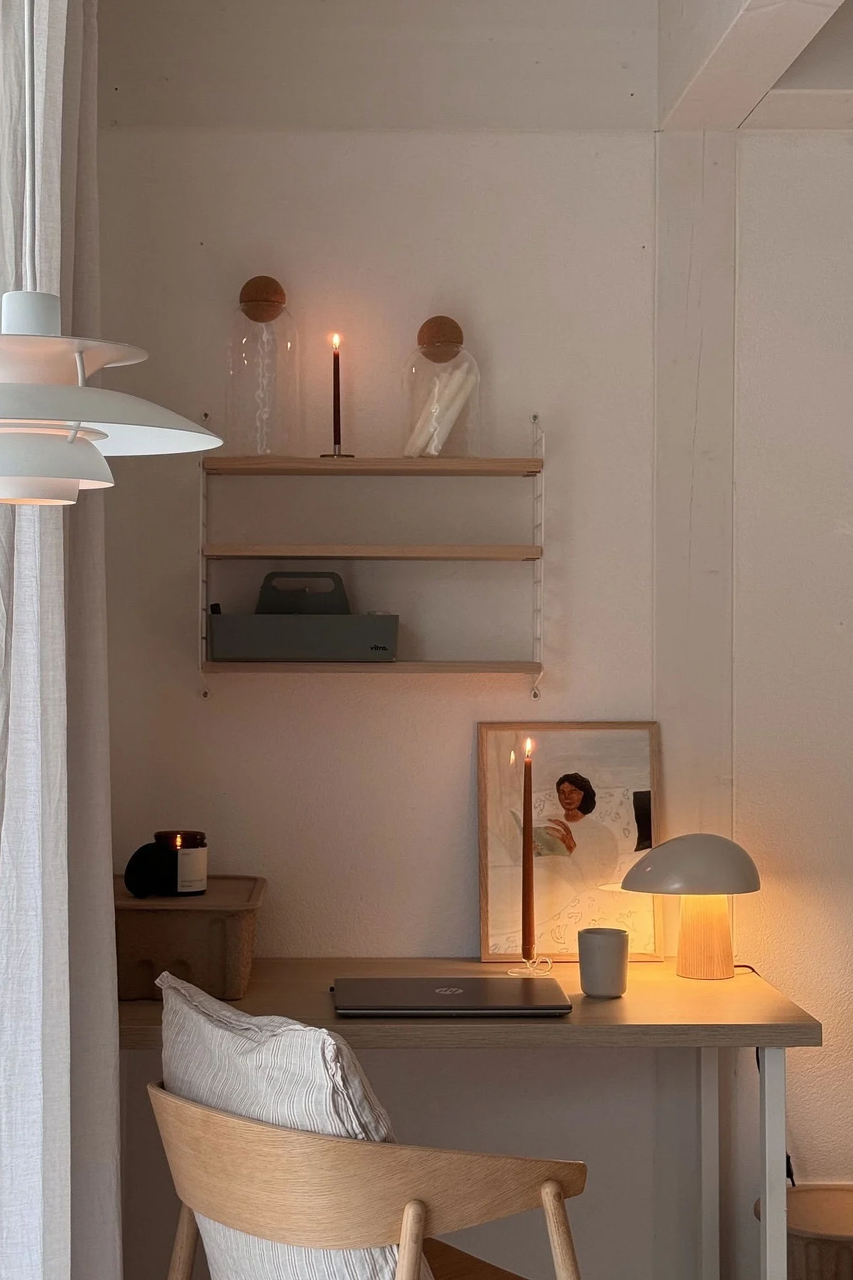 Een gezellige bureau-opstelling met warme verlichting, een gesloten laptop, twee brandende kaarsen, een moderne lamp en een ingelijst kunstwerk.