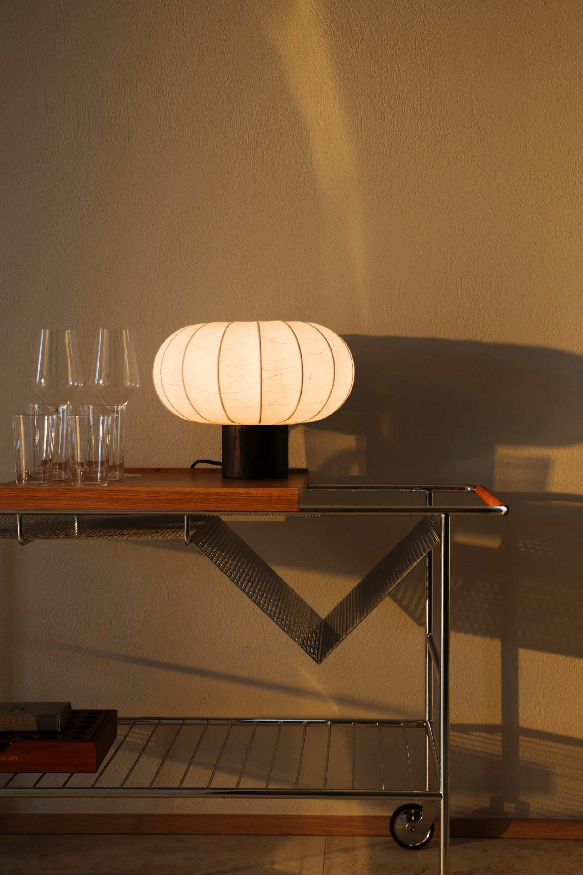 Een gloeiende, geribbelde, ovale lamp staat op een houten en chromen barkar naast heldere glazen, met een gestructureerde muur op de achtergrond.