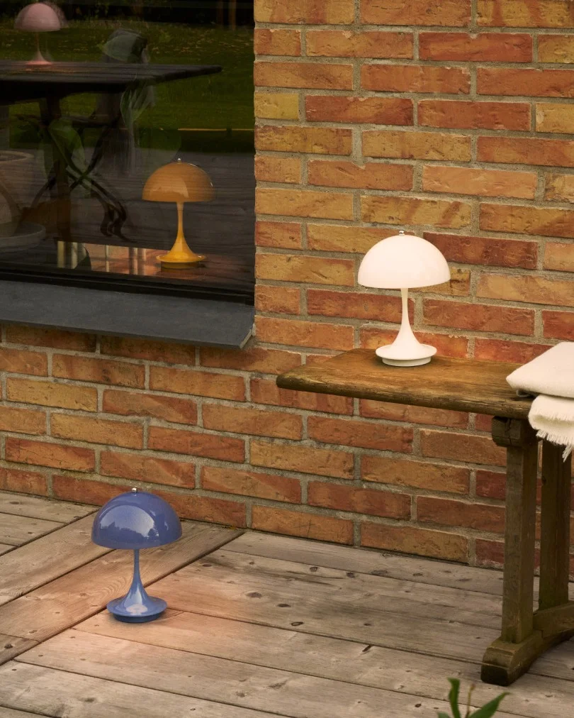 Drie paddenstoelvormige lampen in wit, blauw en geel op een houten terras naast een bakstenen muur en raam.