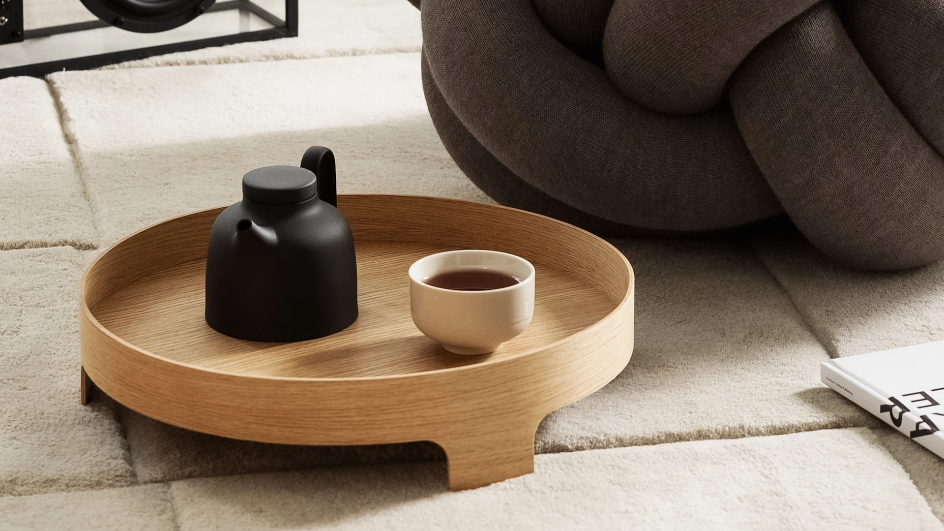 Een zwarte theepot en een kopje thee op een rond houten dienblad, rustend op een beige getextureerd vloerkleed naast een bruin geknoopt kussen.