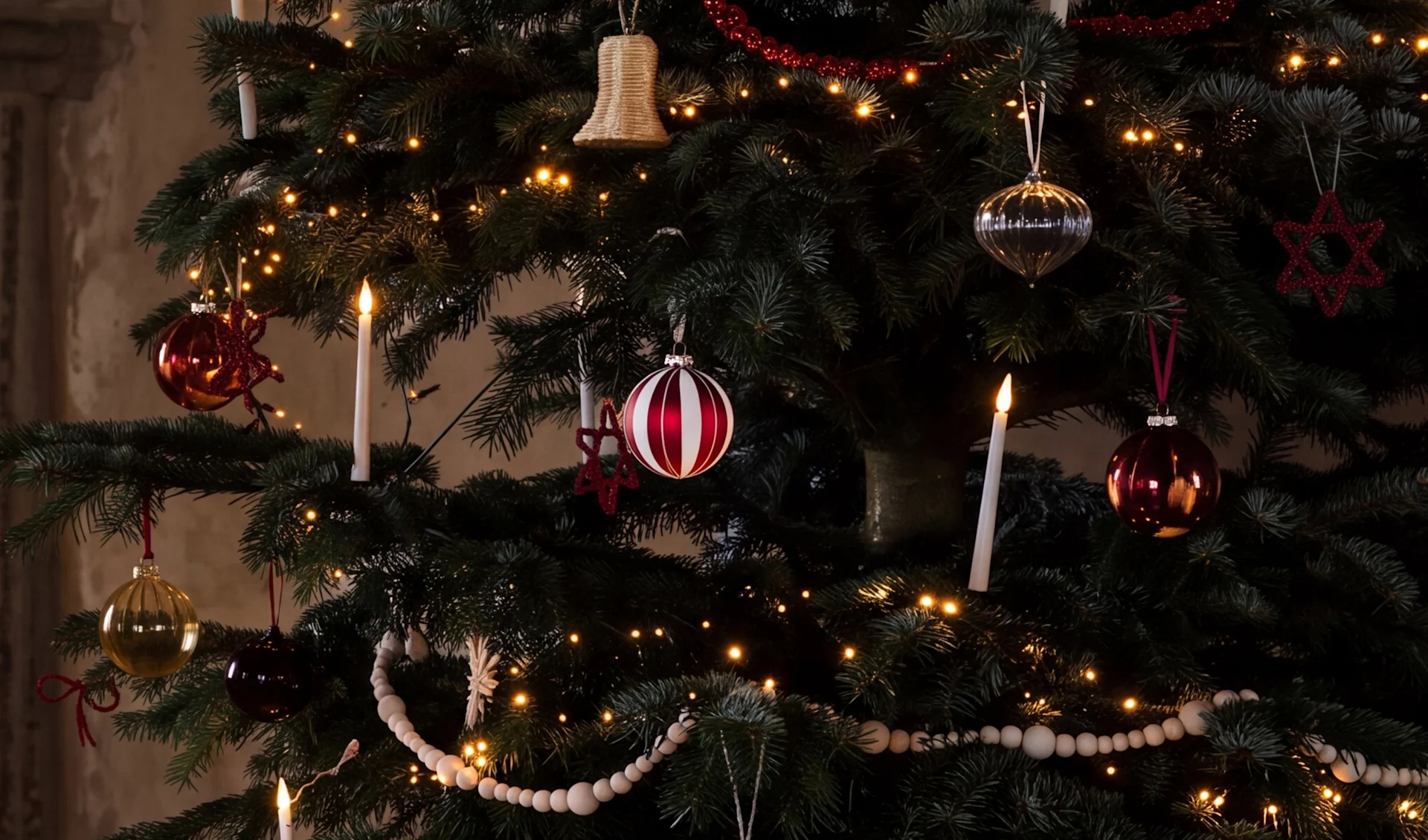 Een versierde kerstboom met warme lichtjes, rode, zilveren en gouden kerstballen, witte kaarsen en houten kralenslingers.