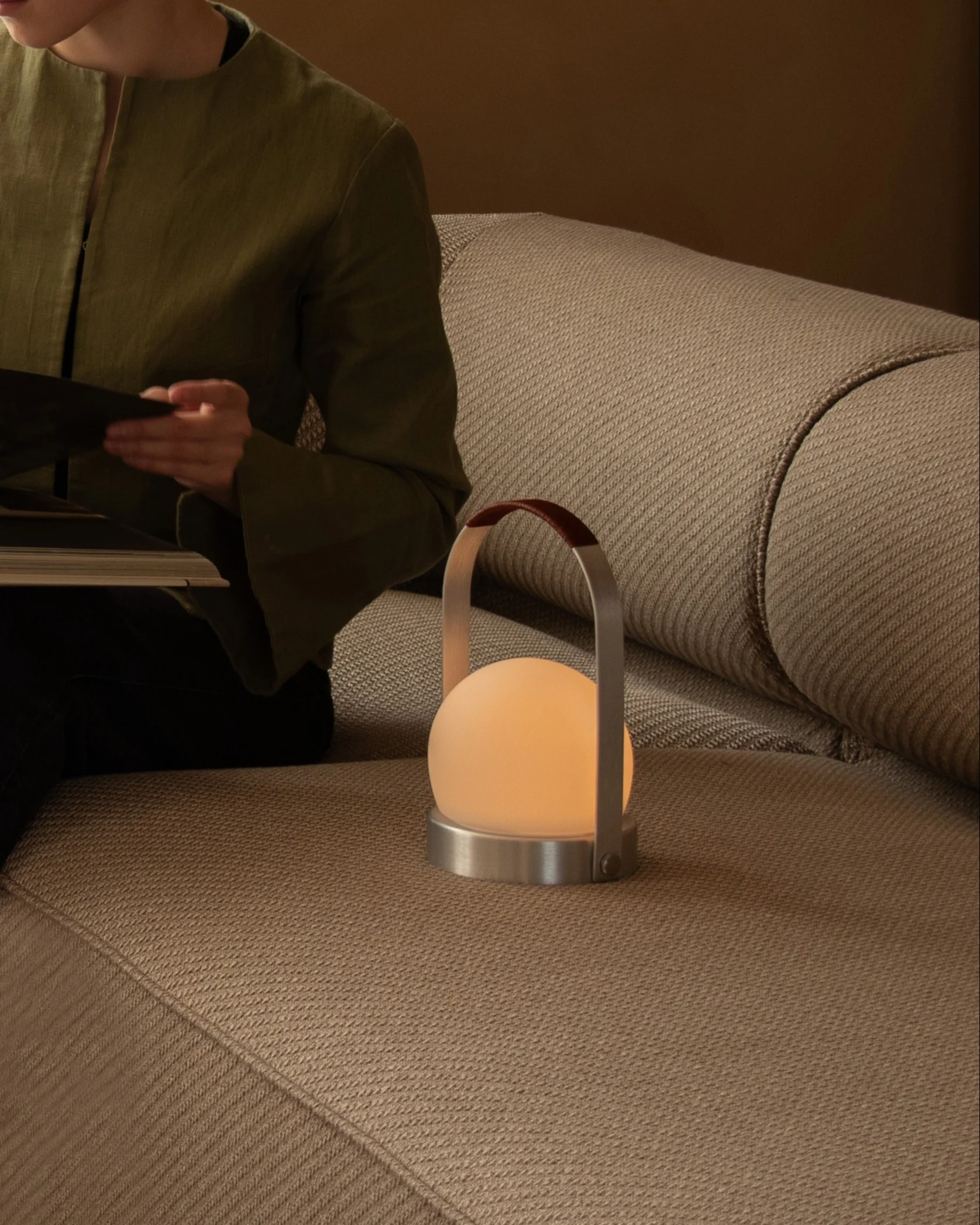 Een persoon leest een boek op een beige bank naast een gloeiende bolvormige draagbare lamp met een leren handvat.