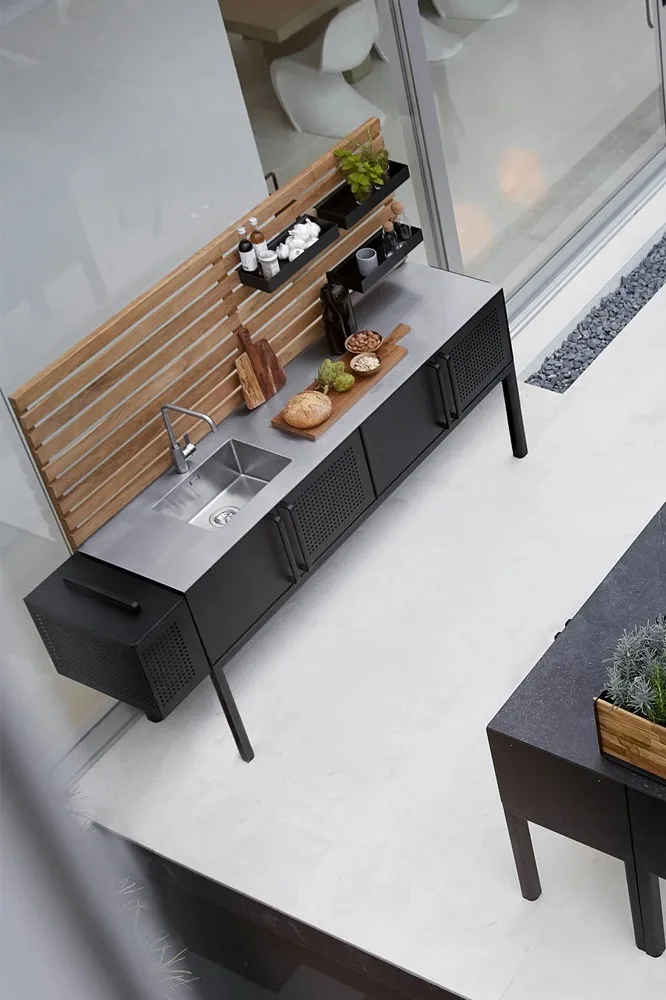 Moderne buitenkeuken met roestvrijstalen spoelbak, zwarte kasten, houten achterpaneel en planken met kruiden en etenswaren.