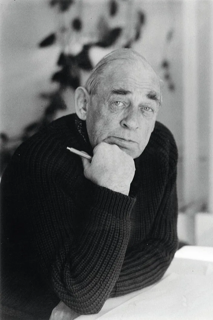 Zwart-wit portret van een oudere man in een donkere trui, die zijn kin op zijn hand laat rusten en een pen vasthoudt, aandachtig naar de kijker kijkend.