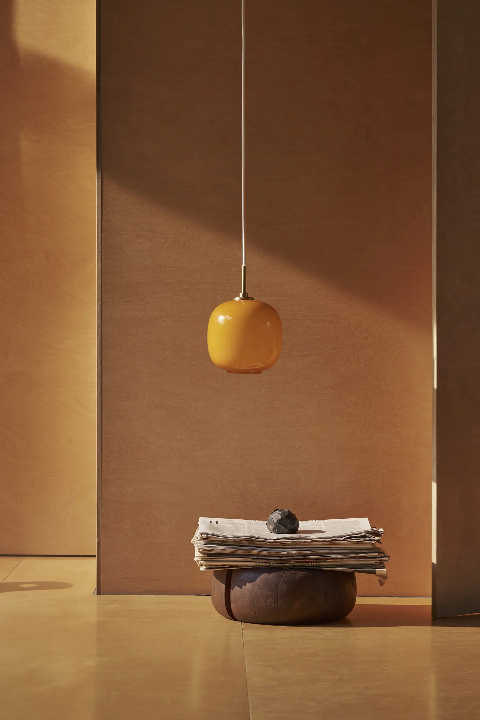 Een oranje ovale hanglamp hangt boven een stapel kranten en een donker decoratief object op een houten onderstel in een minimalistische kamer.