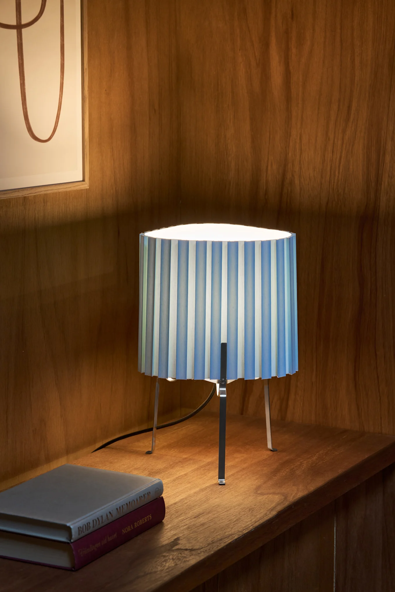 Een tafellamp met een blauw-wit gestreepte geplooide kap en metalen poten, staand op een houten plank naast twee boeken.