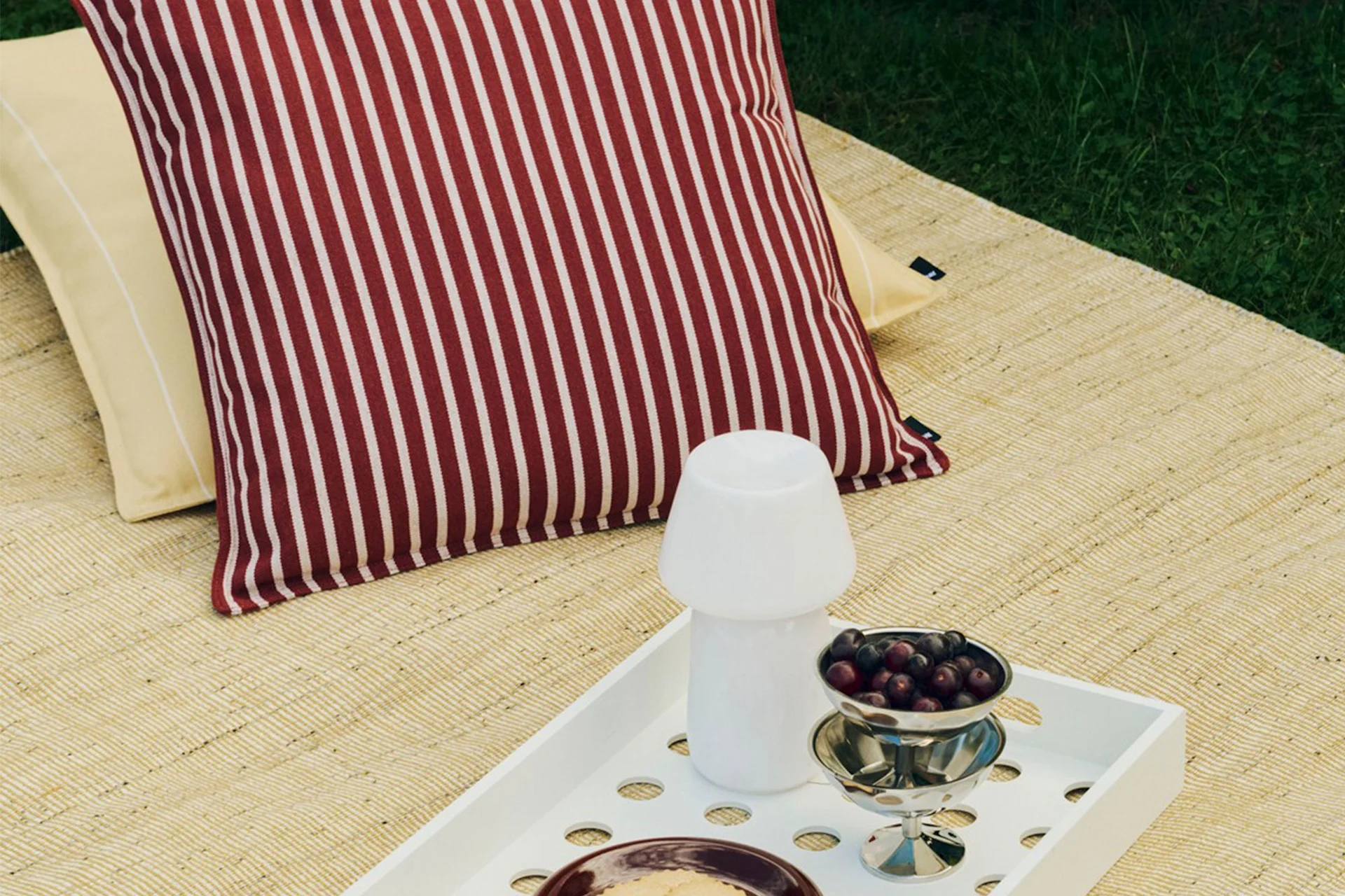 Buiten picknickopstelling met gestreept rood-wit kussen, geel kussen, wit dienblad met paddenstoellamp en een kom bessen.