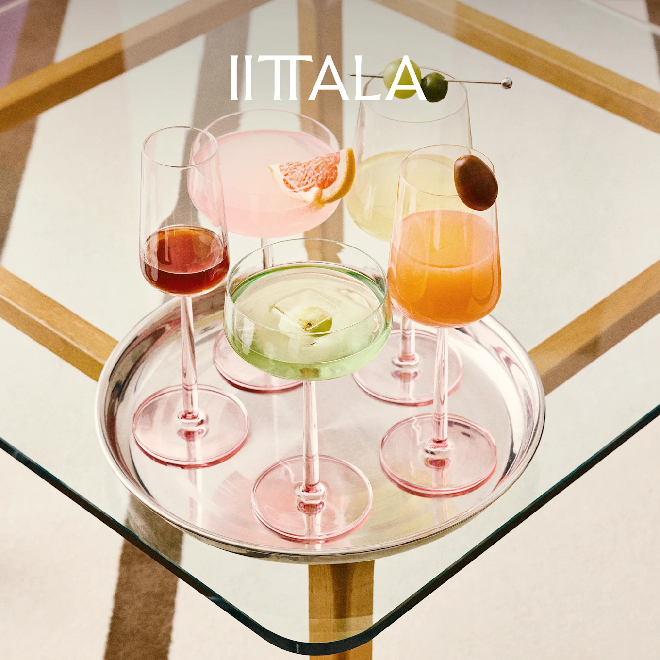 IITTALA-logo boven een zilveren dienblad met vijf kleurrijke cocktails in roze-gestemde glazen op een glazen tafel.