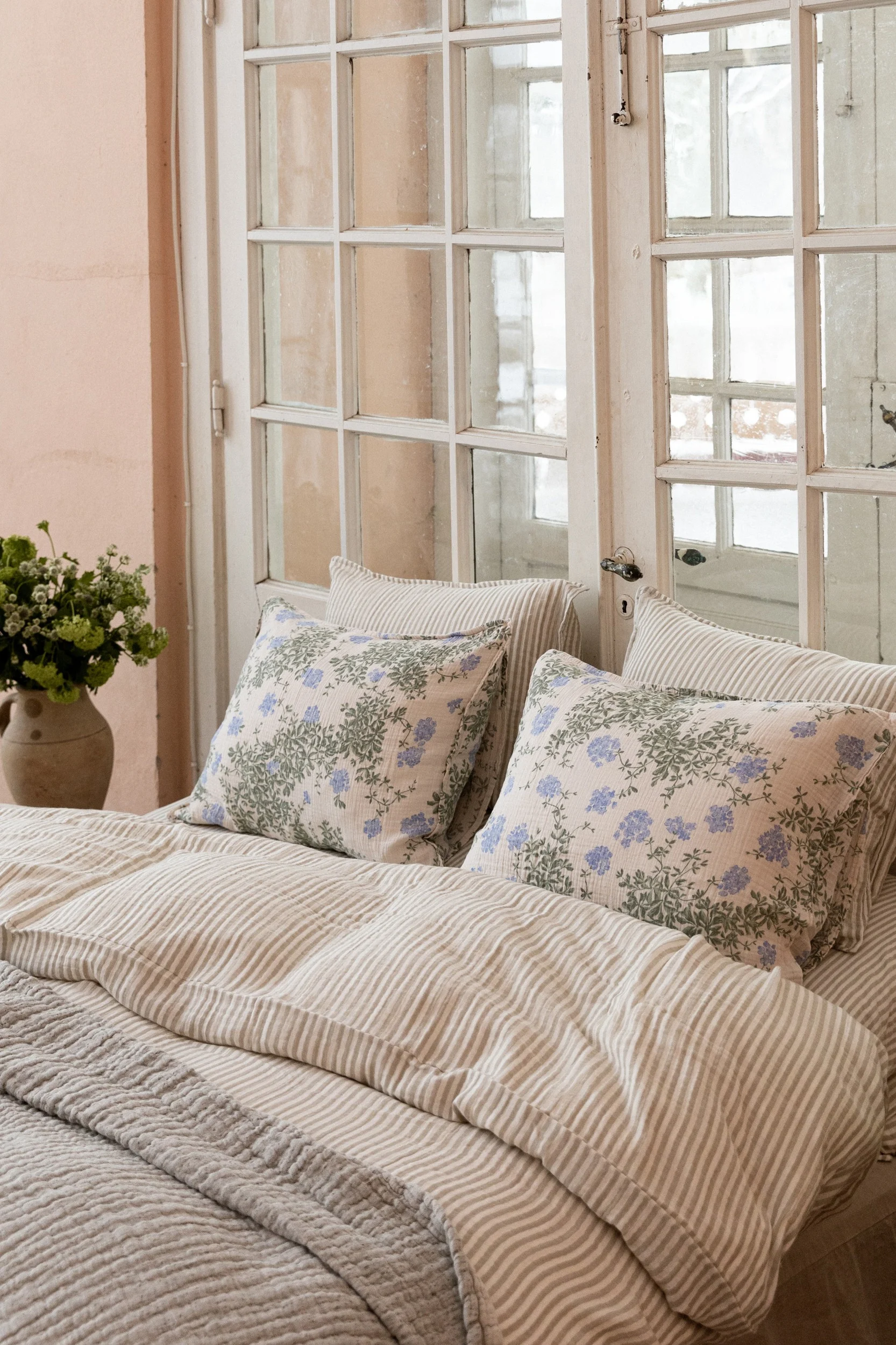 Een bed met een gestreept dekbed en twee kussens met blauw bloemenpatroon, voor een raam met panelen. Een vaas met groen staat links.