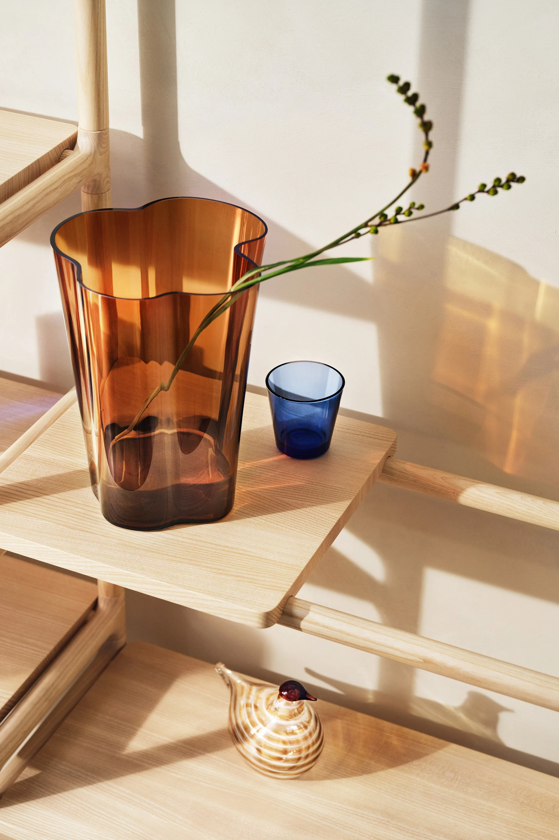 Amberkleurige vaas met tak, blauwe beker en gestreepte glazen vogel op lichte houten plankeneenheid.