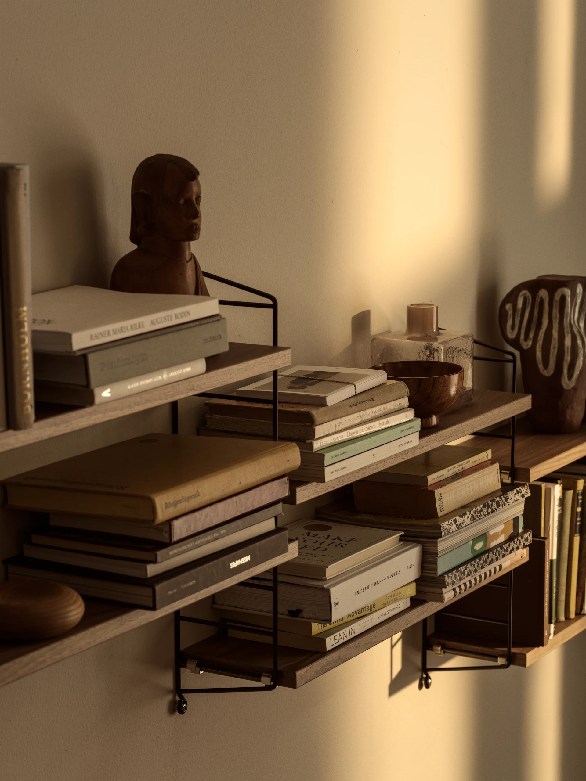 Wandplanken gevuld met boeken, een houten buste en decoratieve objecten, verlicht door warm zonlicht op de muur.