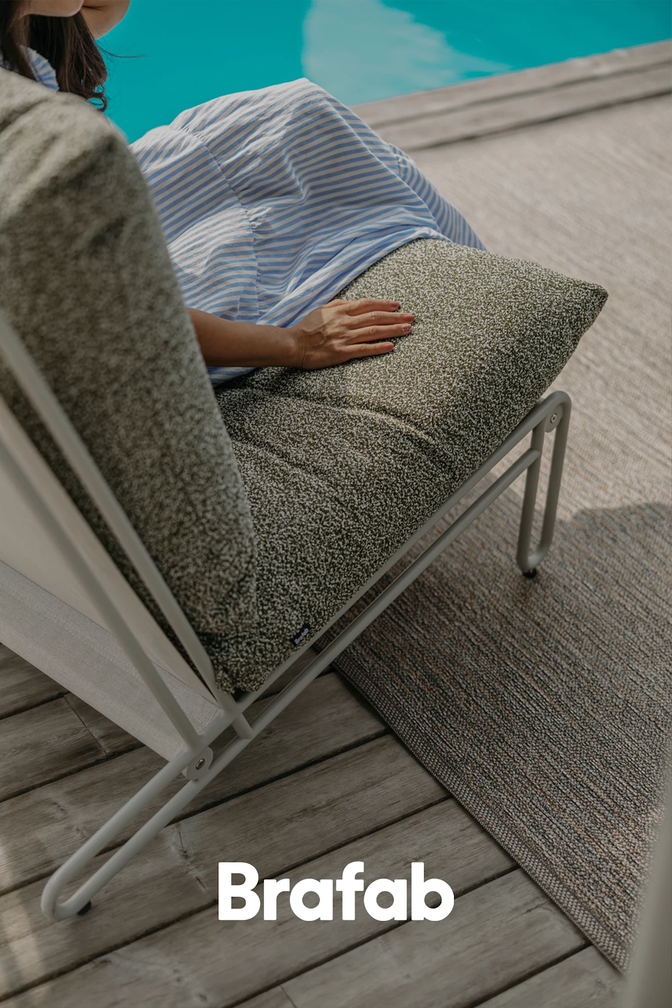 Persoon zit op een moderne outdoor loungefauteuil met donkergroen gespikkeld kussen op een houten terras bij een zwembad.