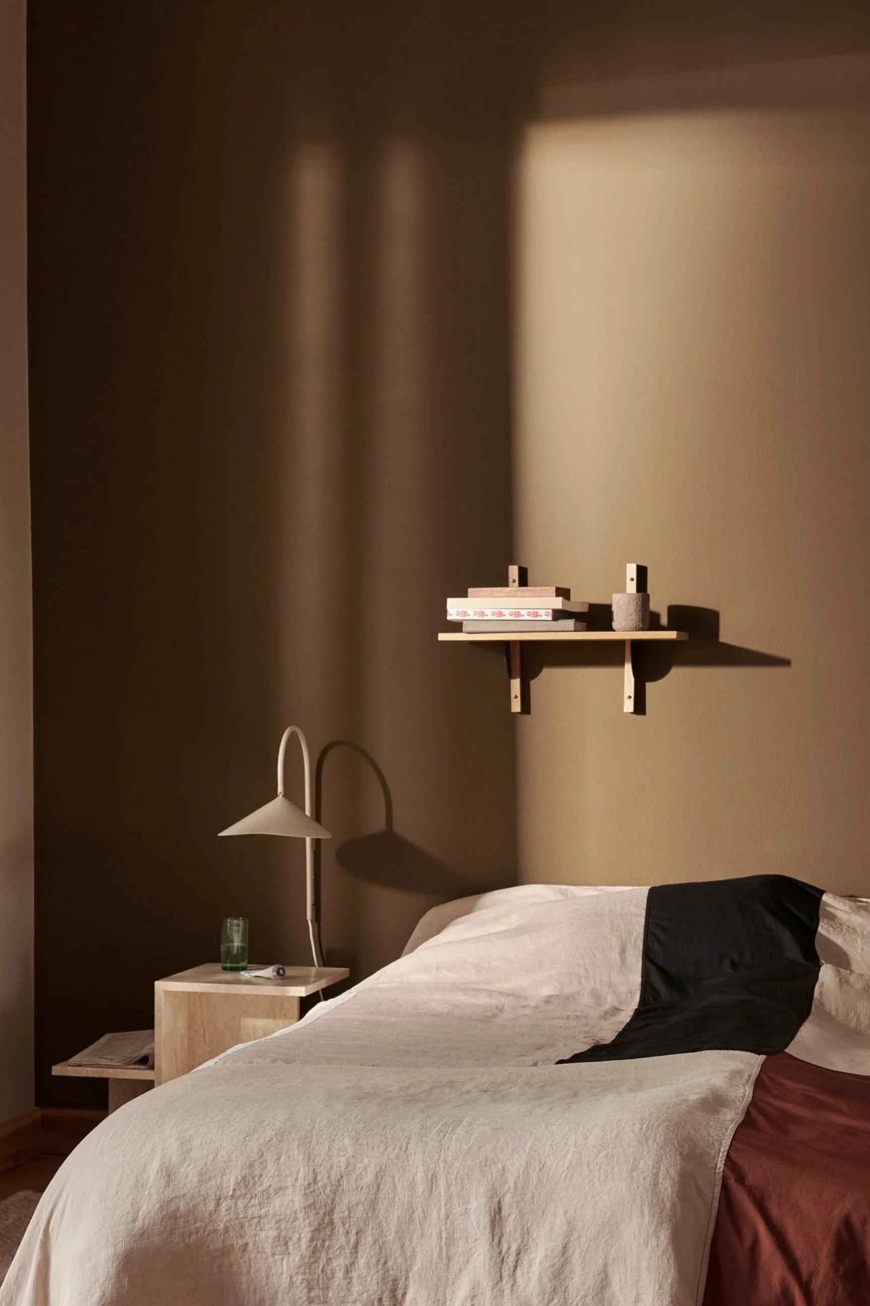 Een slaapkamer met een bed met beige, zwart en roestkleurig beddengoed, een nachtkastje met lamp en een plank met boeken aan een warmbruine muur.