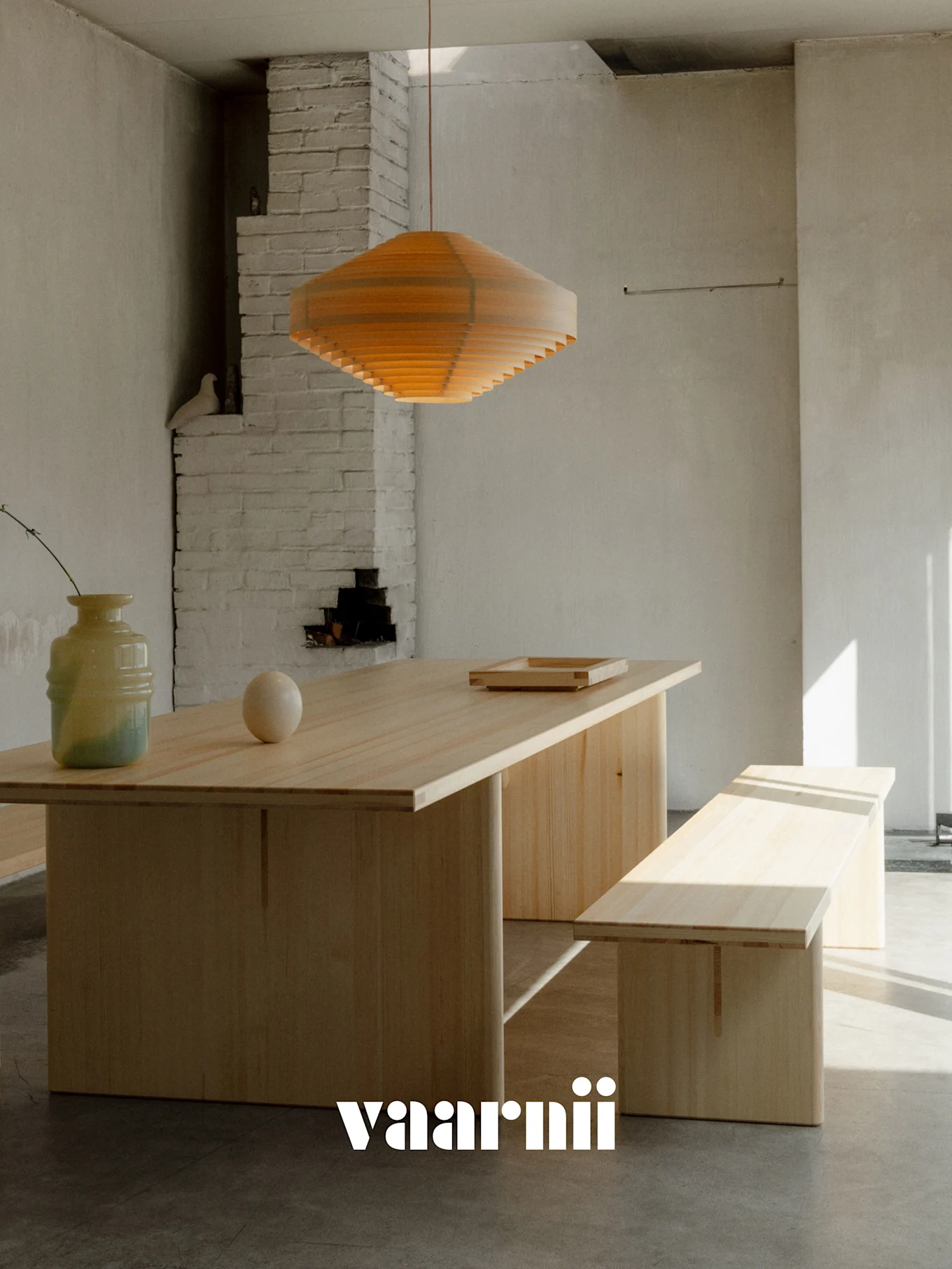 Een minimalistische eetkamer met een lange lichte houten tafel, bijpassende bank, gelaagde houten hanglamp, vaas en bol. Vaarnii-logo.