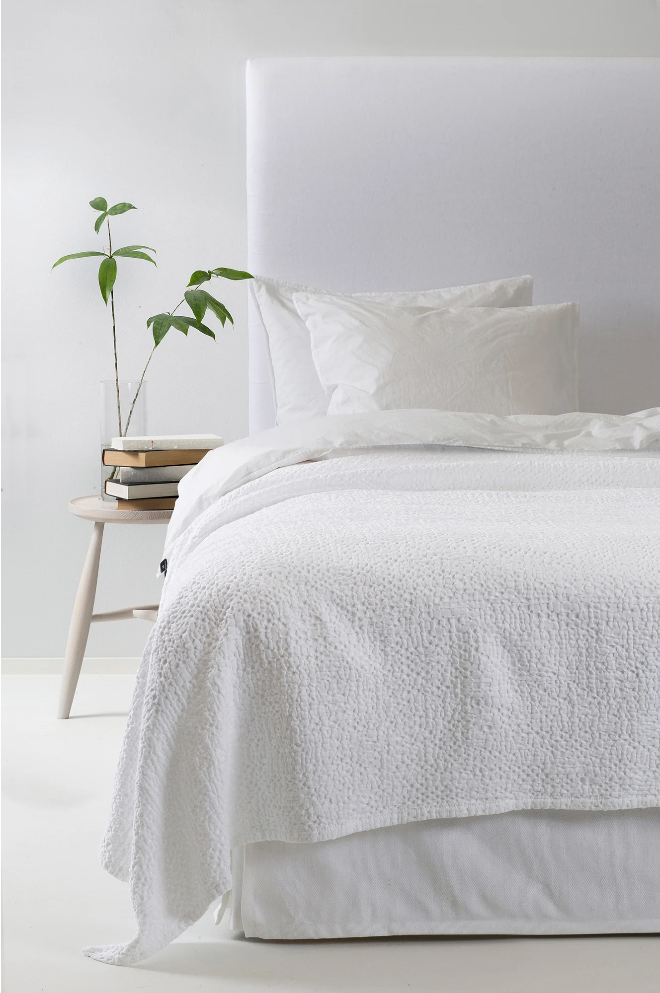 Wit bed met getextureerd dekbed, kussens, gestoffeerd hoofdbord, houten bijzettafel met boeken en groene plant.