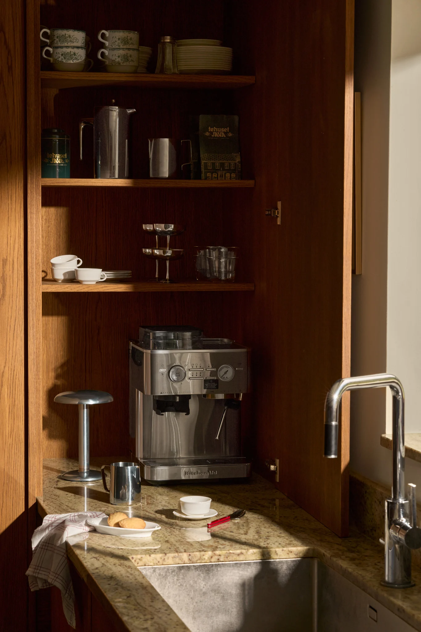 Een open houten keukenkast met een espressomachine, koffieaccessoires, gestapelde mokken en glazen op planken, naast een gootsteen.