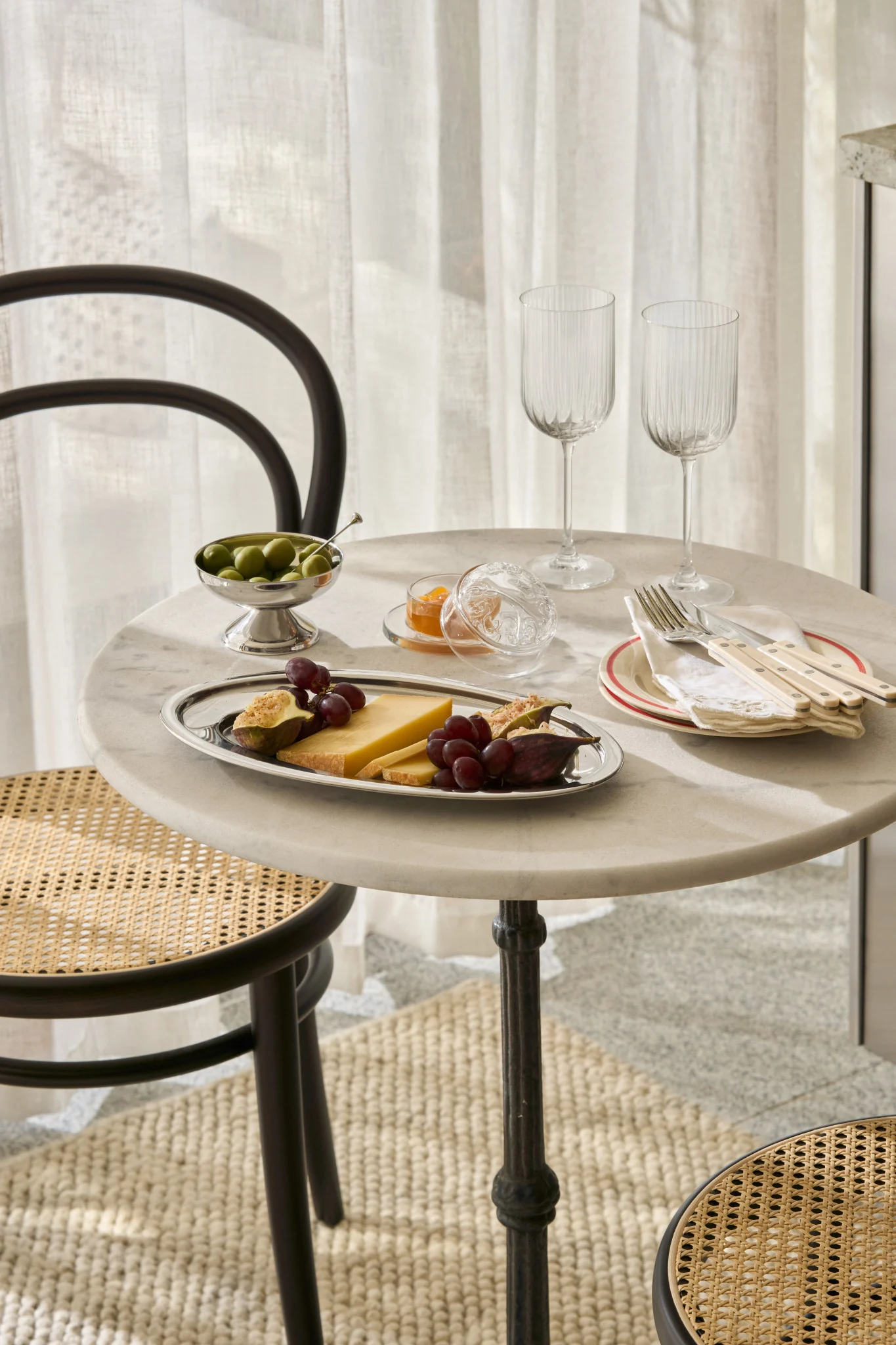 Een marmeren tafel met kaas, vijgen, druiven, olijven, jam, glazen en bestek, met rotan stoelen.