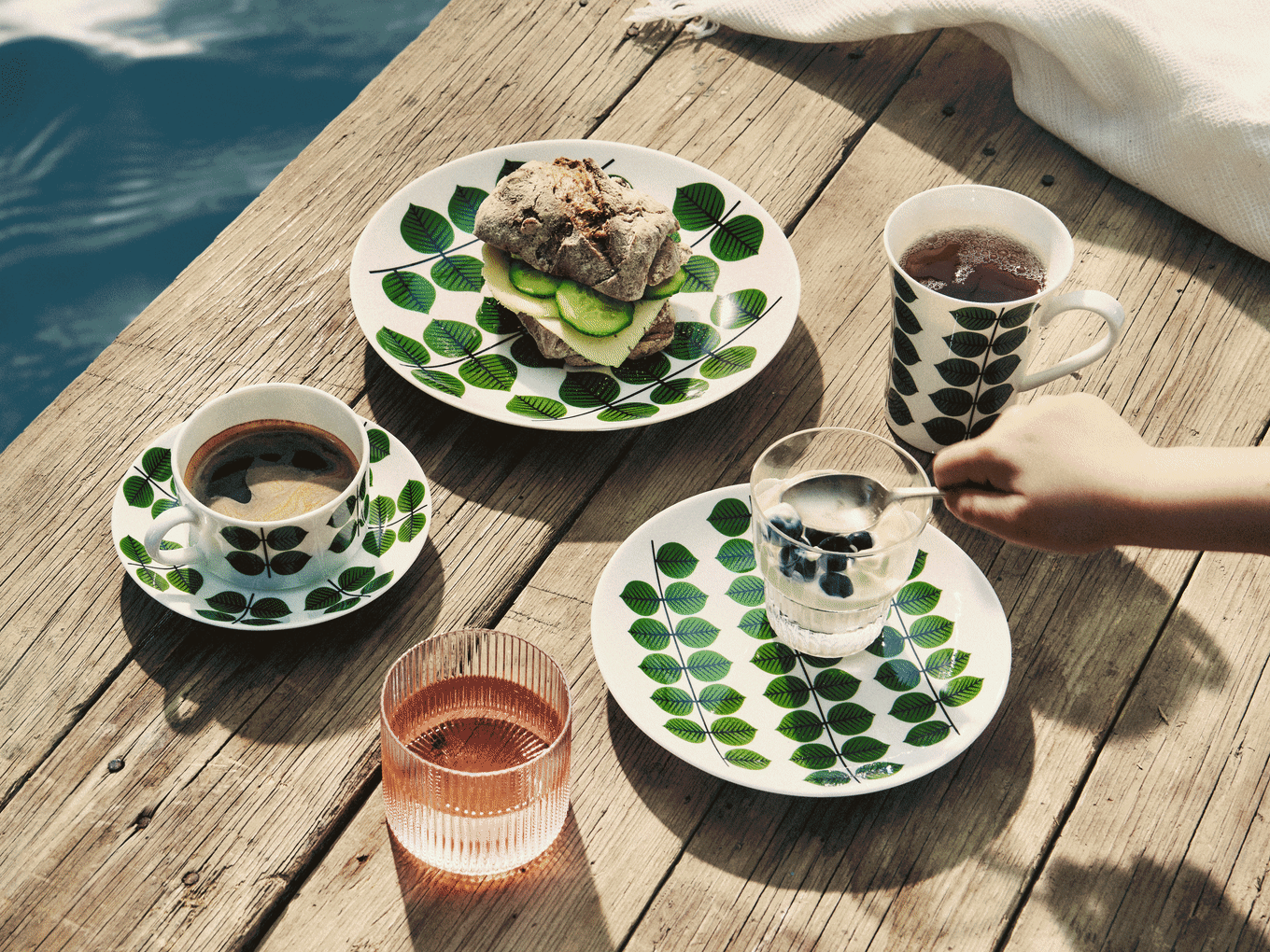 Ontbijt op houten pier: Bladmotief servies met sandwich, koffie, thee, yoghurt met bosbessen en roze drankje.