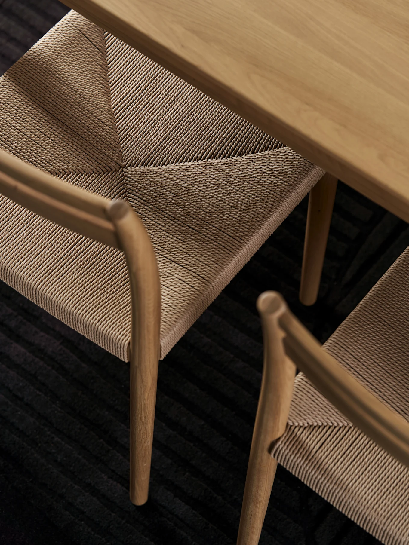 Bovenaanzicht van een lichte houten tafel en twee stoelen met gevlochten zittingen op een donker geribbeld vloerkleed.