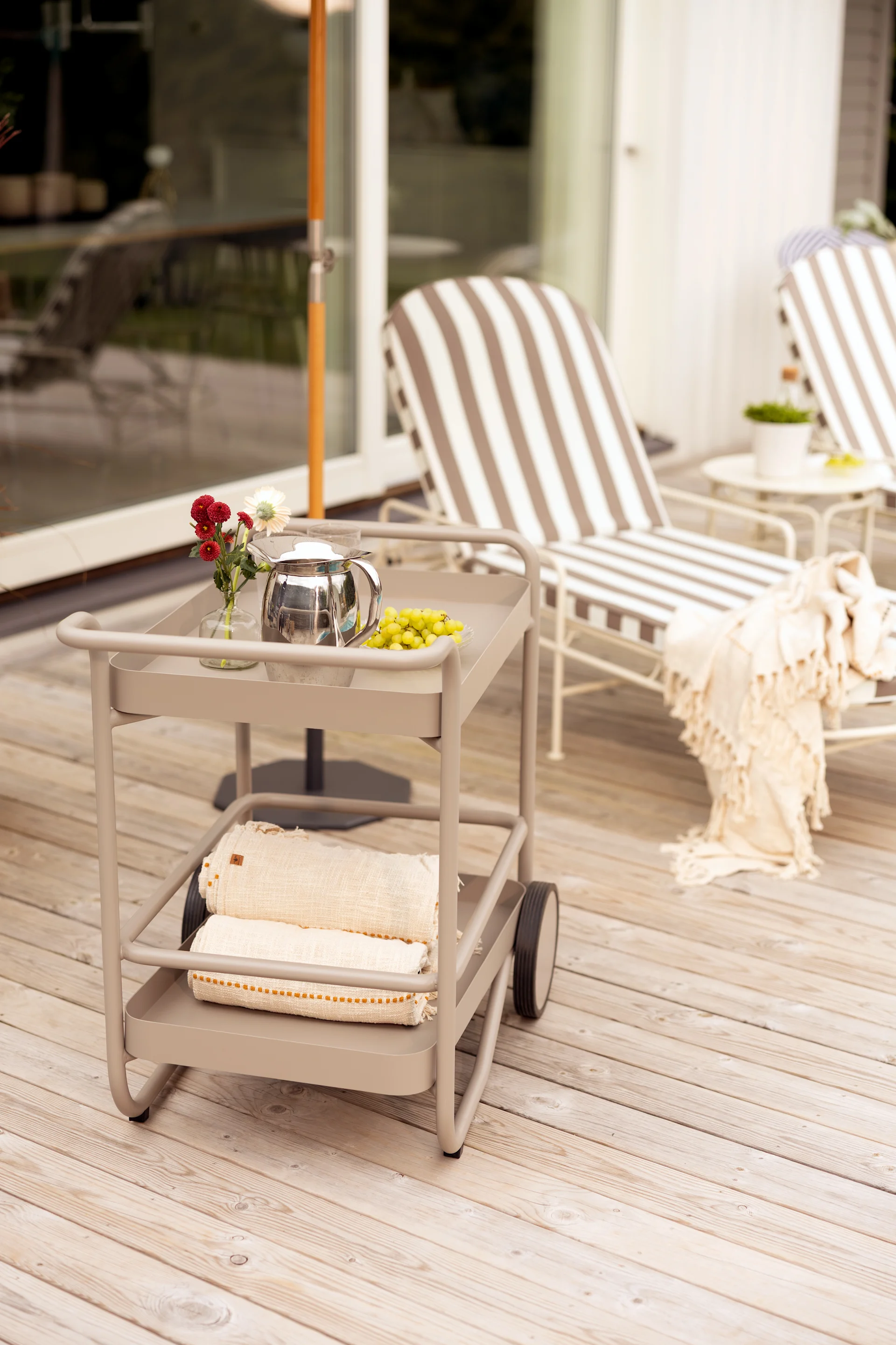 Twee-laags beige buiten serveerwagen met theepot, druiven en bloemen bovenop, opgerolde handdoeken onderaan.