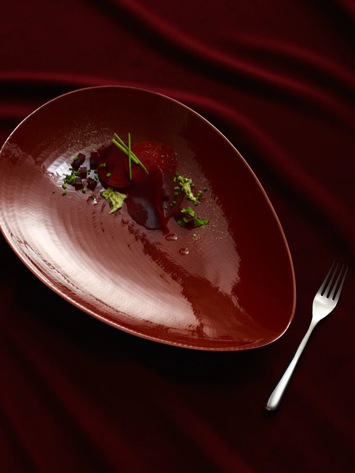 Een donkerrood peervormig bord met een bieten gerecht, gegarneerd met groene kruiden en donkere blokjes, naast een zilveren vork op een gedrapeerde donkerrode stof.