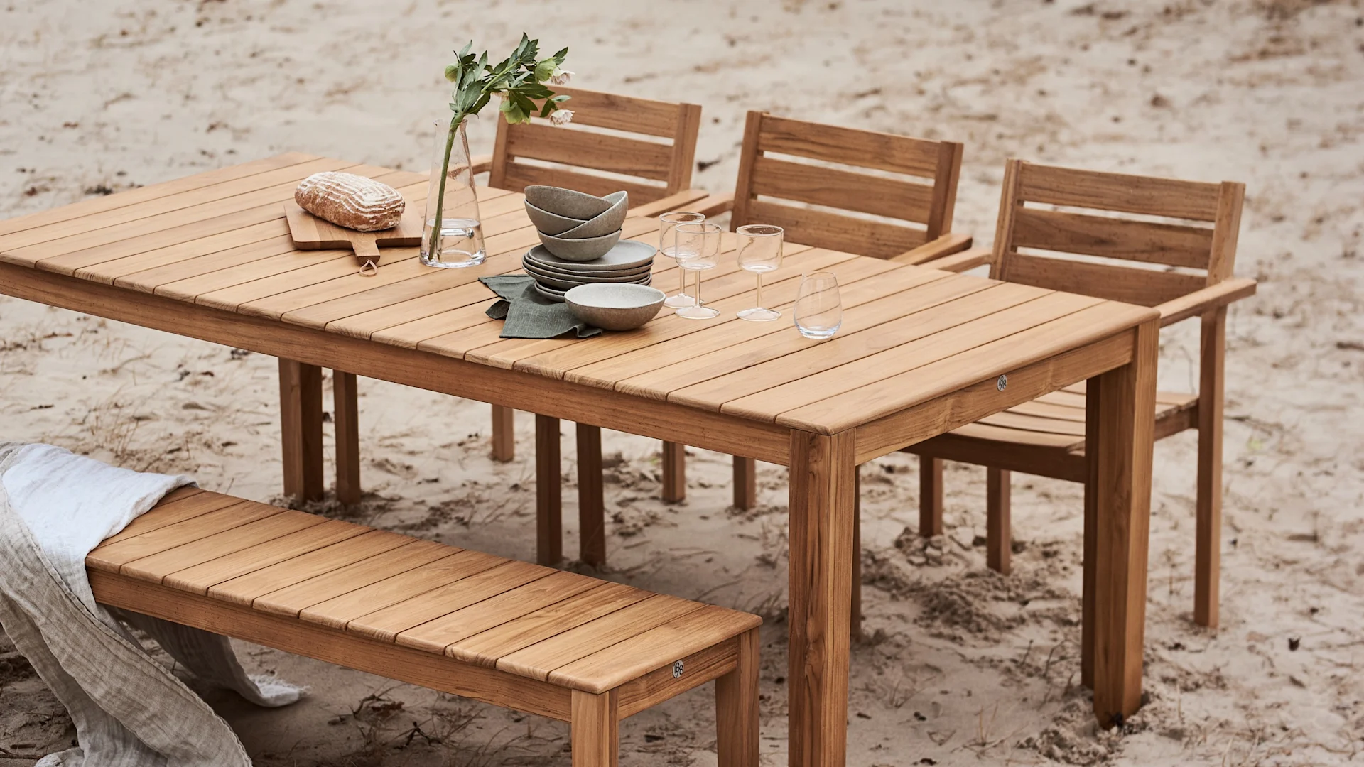 Houten buiteneettafel gedekt met servies, glazen, een vaas met bloemen en brood, omringd door stoelen en een bank op zand.