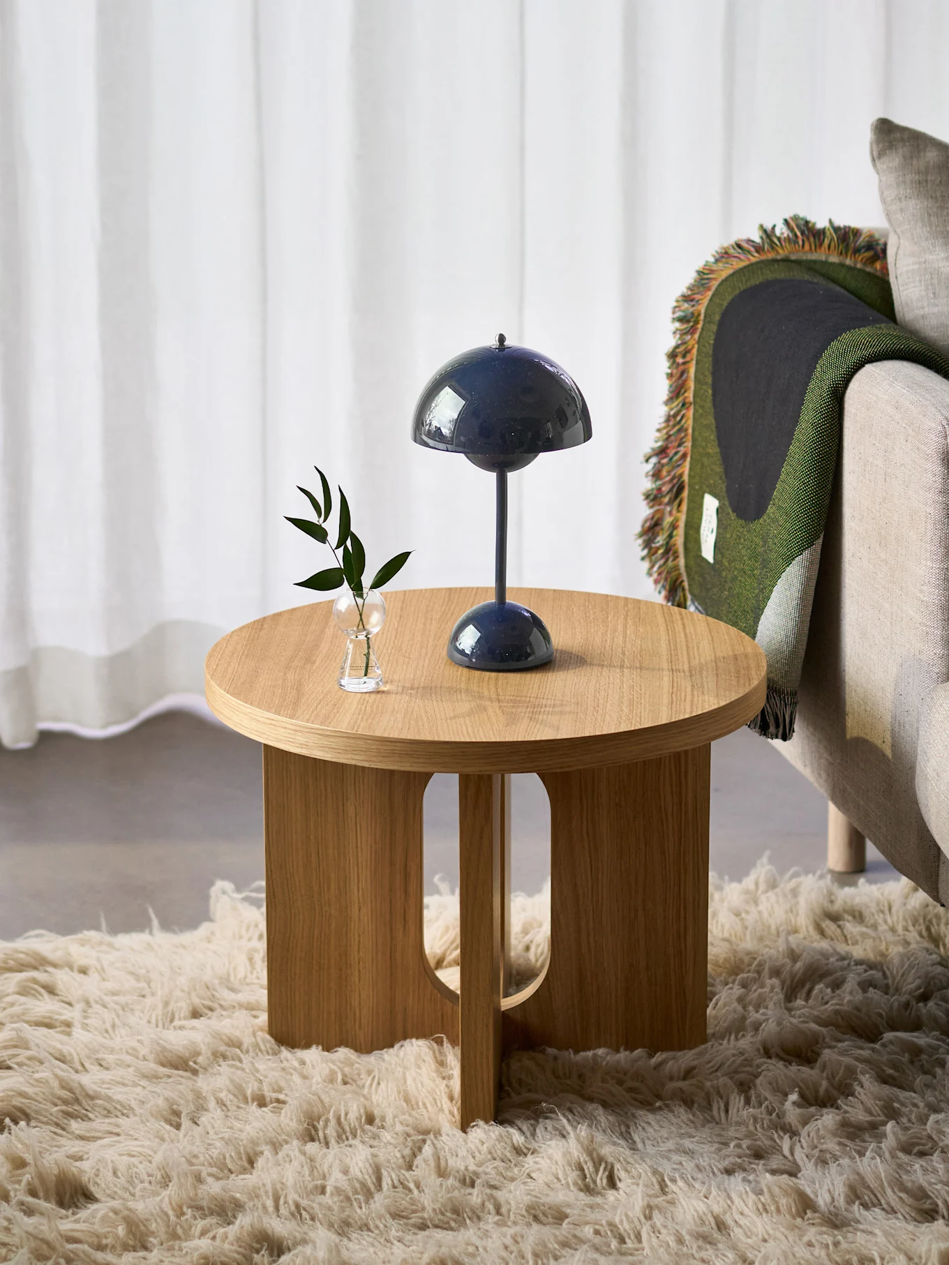 Rond houten bijzettafeltje met blauwe lamp en plant, naast fauteuil met groene deken, op een pluizig vloerkleed.