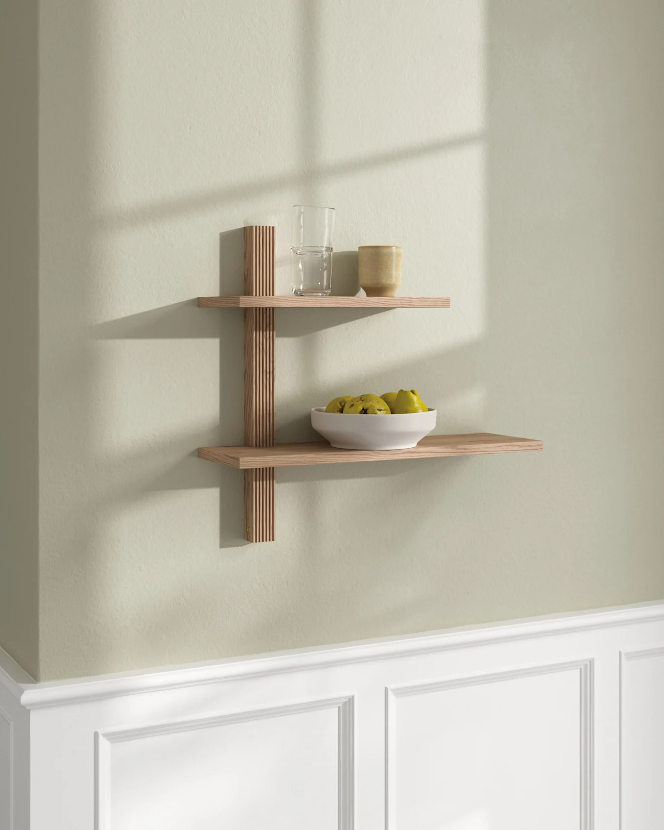 Twee lichte houten zwevende planken aan een lichtgroene muur. Bovenste plank met glas en mok; onderste plank met witte fruitschaal.