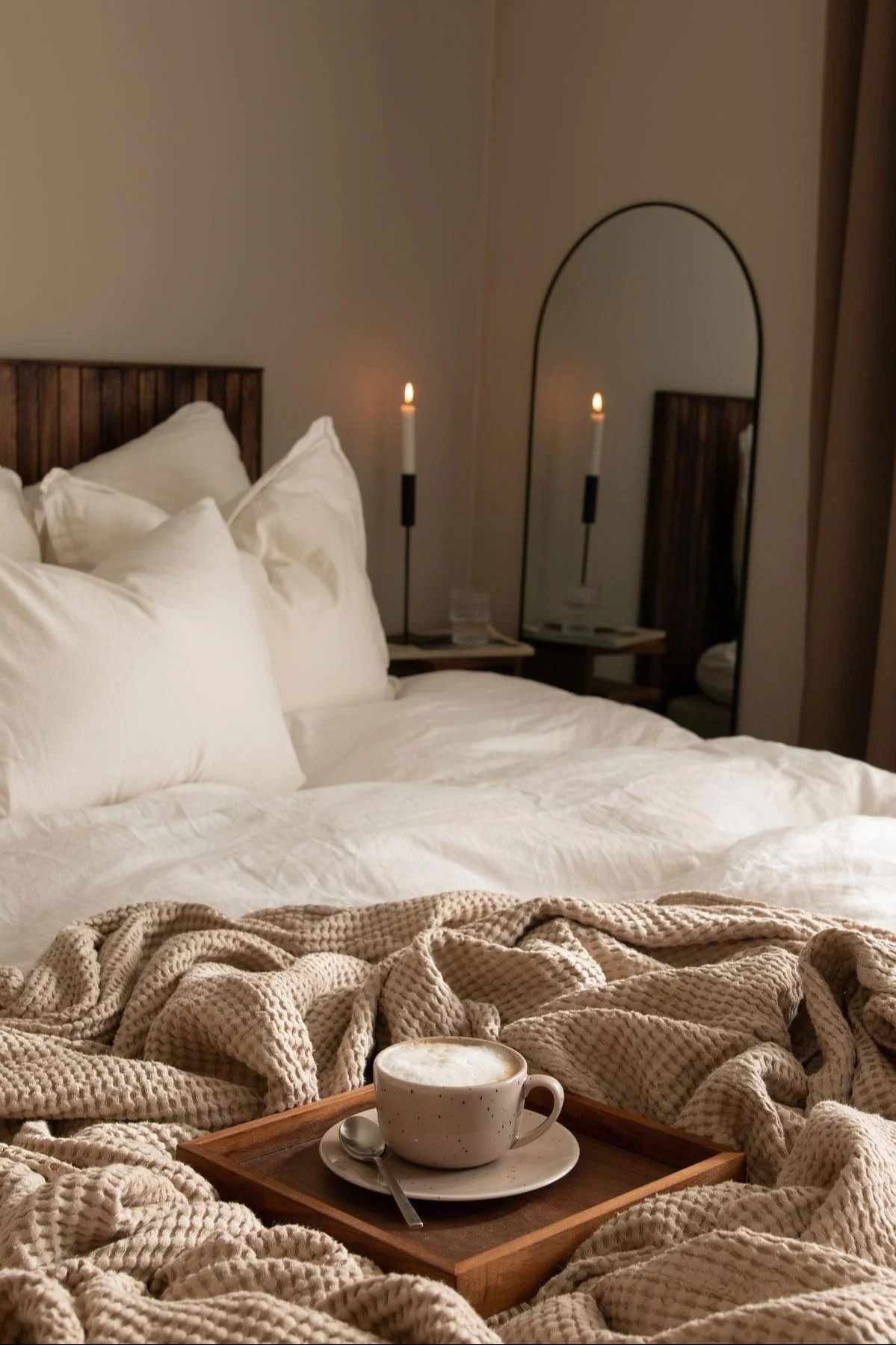 Een knusse slaapkamer met een wit bed, beige gebreide plaid en een kop koffie op een houten dienblad. Branded kaarsen en een spiegel zijn op de achtergrond.