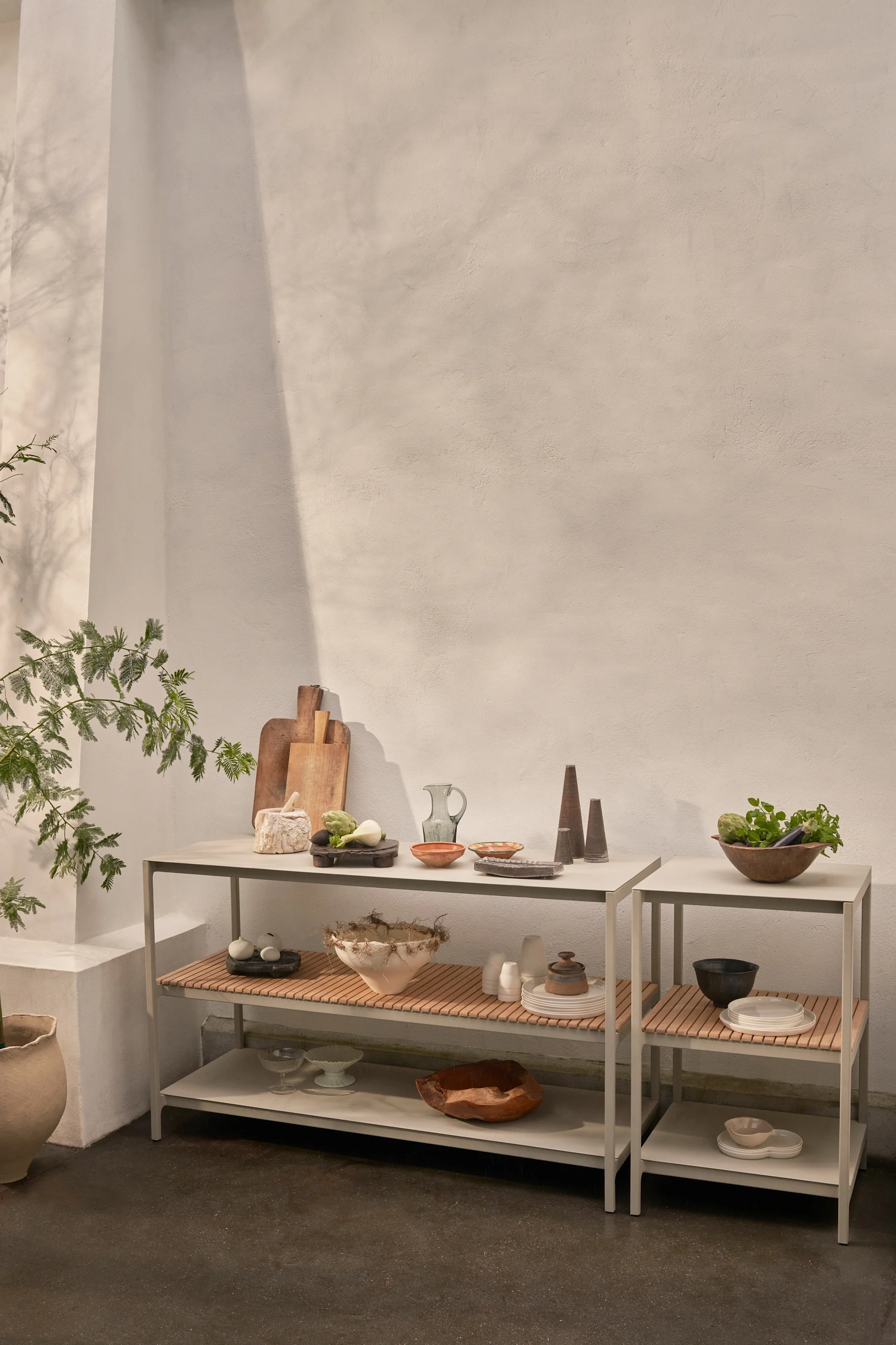 Twee moderne grijze rekken met houten lattenbladen tonen diverse keukenartikelen, keramische schalen, verse groenten en decoratieve objecten tegen een lichte muur.