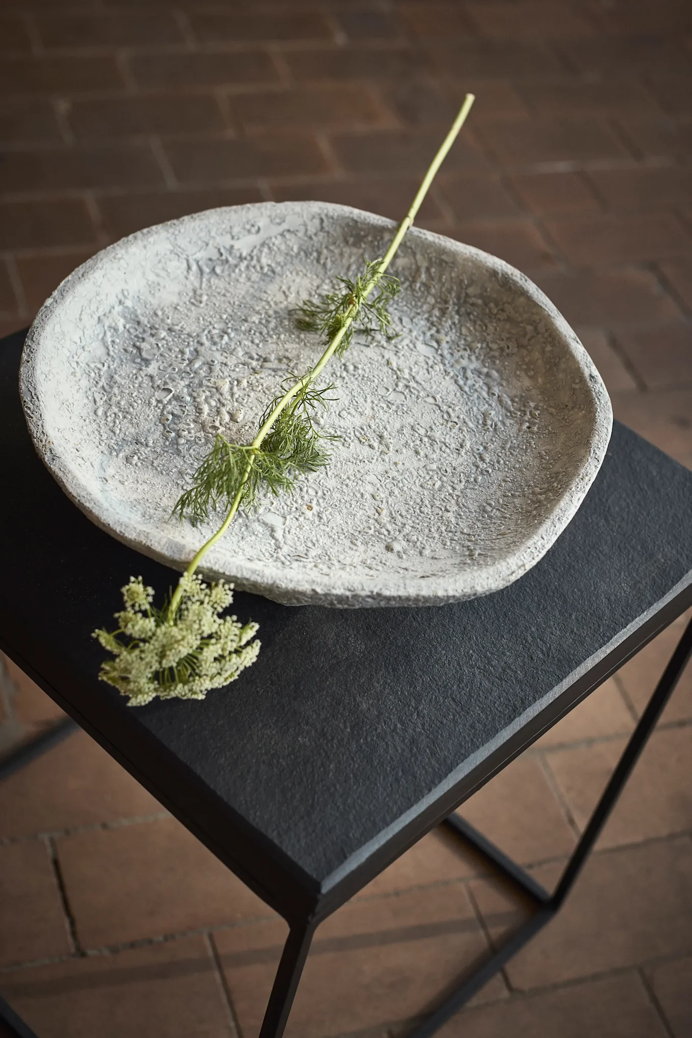Grijze kom met textuur op een zwarte tafel met een groene steel en kleine witte bloemen.