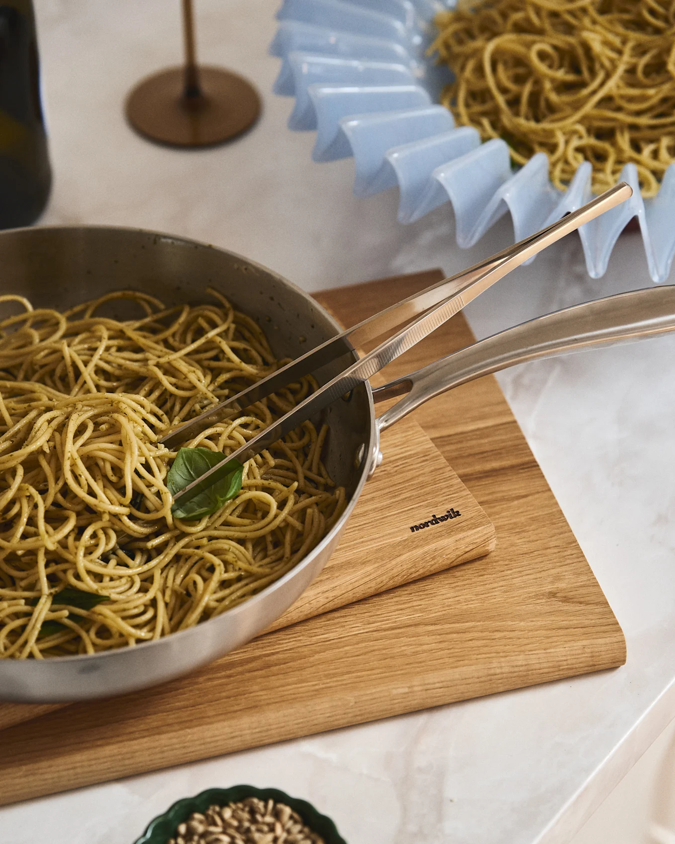 Pesto spaghetti in een zilveren pan met tang op een Nordwik houten plank. Nog een bord spaghetti in een lichtblauwe gegolfde kom.