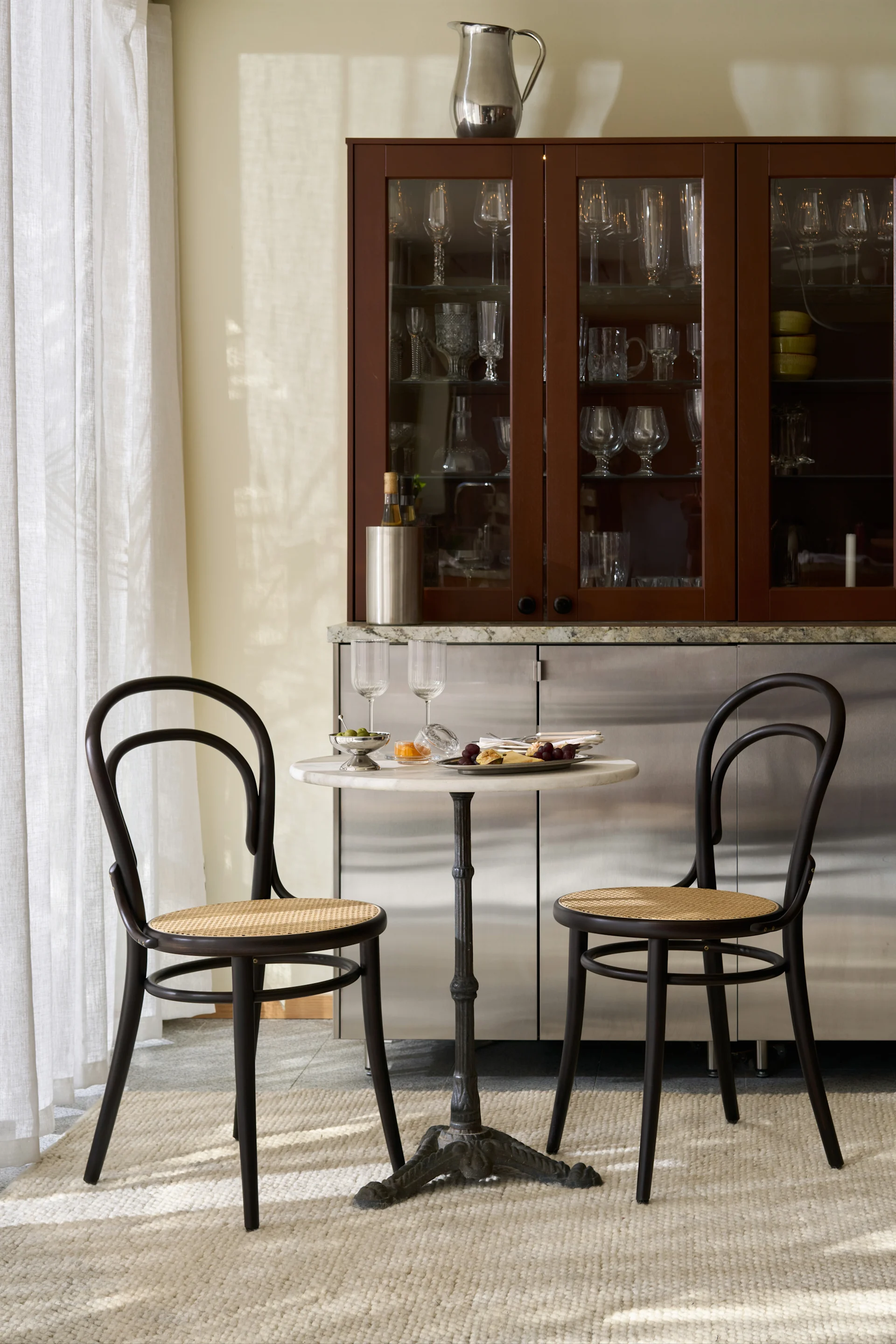 Een stijlvolle eethoek met een marmeren bistrotafel, twee donkerhouten stoelen en een barkast vol glaswerk.
