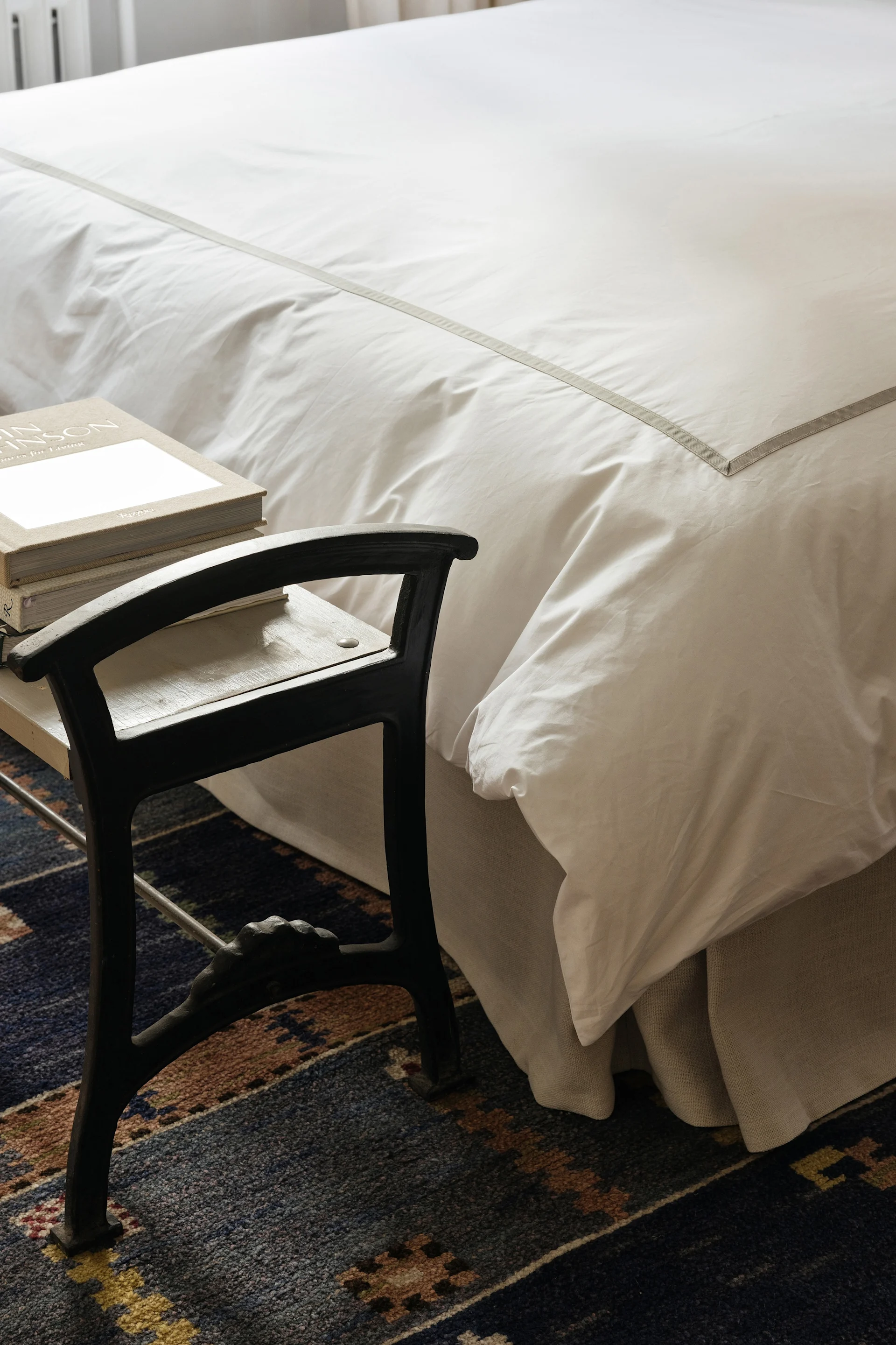 Een bed met wit beddengoed en een grijze streep naast een donkere kruk met boeken op een gedessineerd blauw vloerkleed.